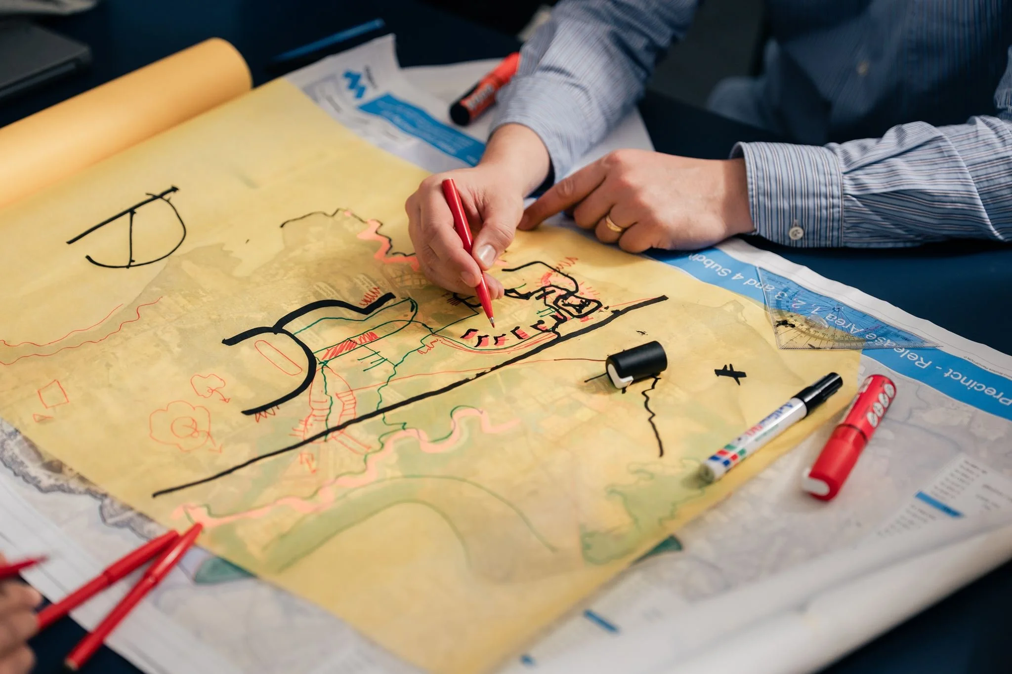 Person drawing on a large map with colored markers, surrounded by markers, placed on a desk.