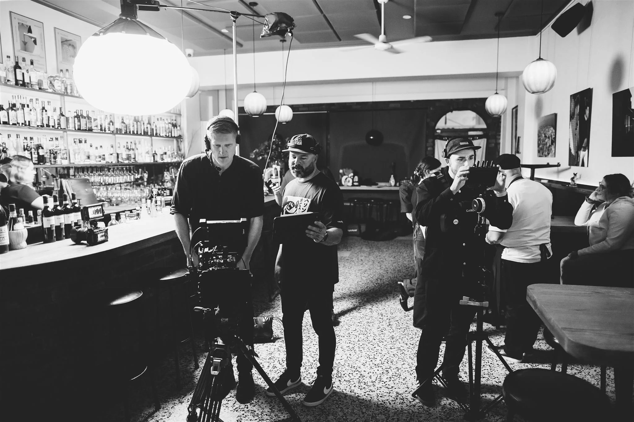 Black and white photo of a film crew filming inside a restaurant or bar. Two men are working with camera equipment in the foreground, with shelves of bottles behind the bar and tables with people in the background.