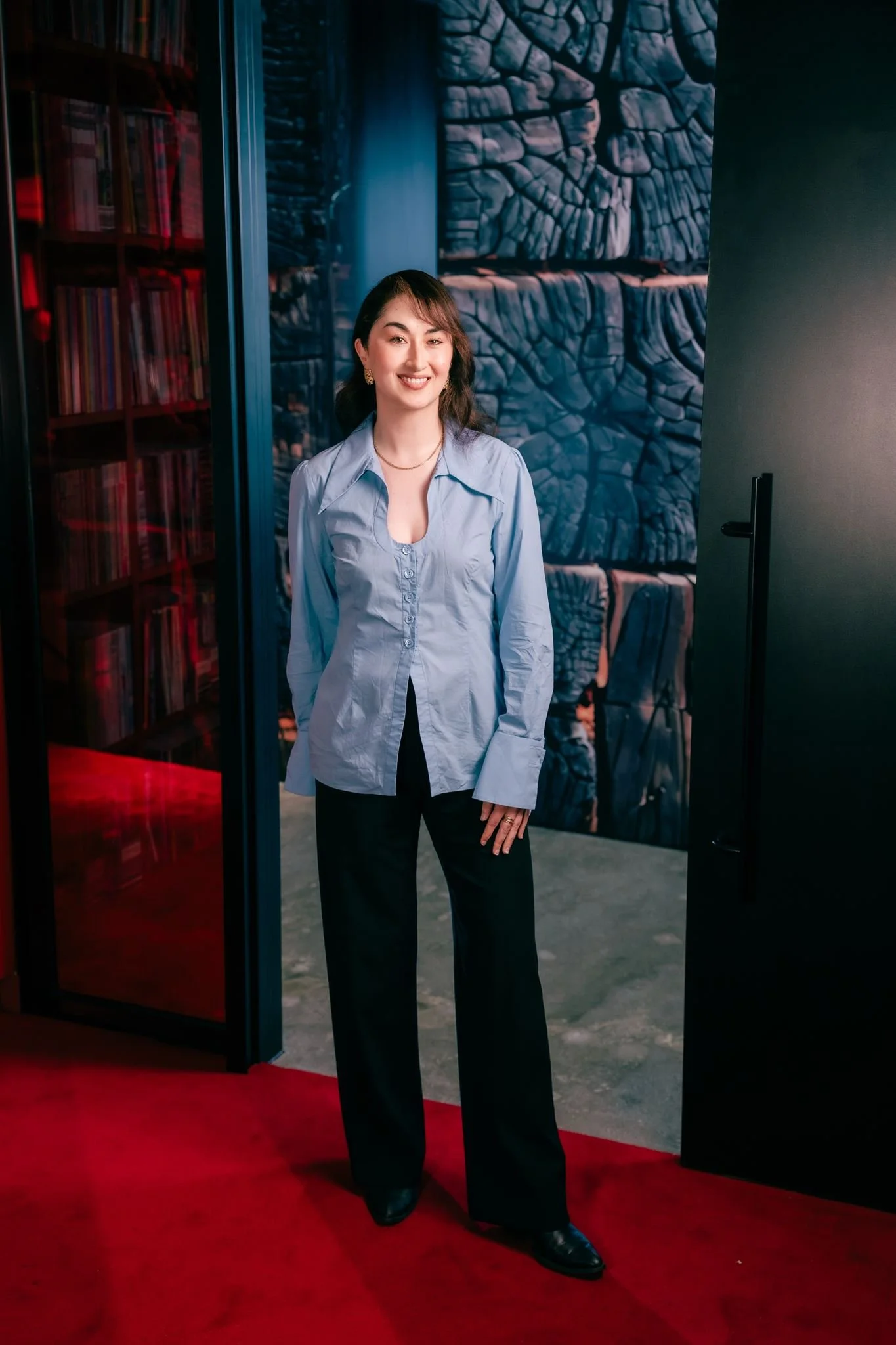 A woman standing on a red carpet, smiling, in front of a textured blue wall, wearing a light blue button-up shirt, black pants, and black shoes.