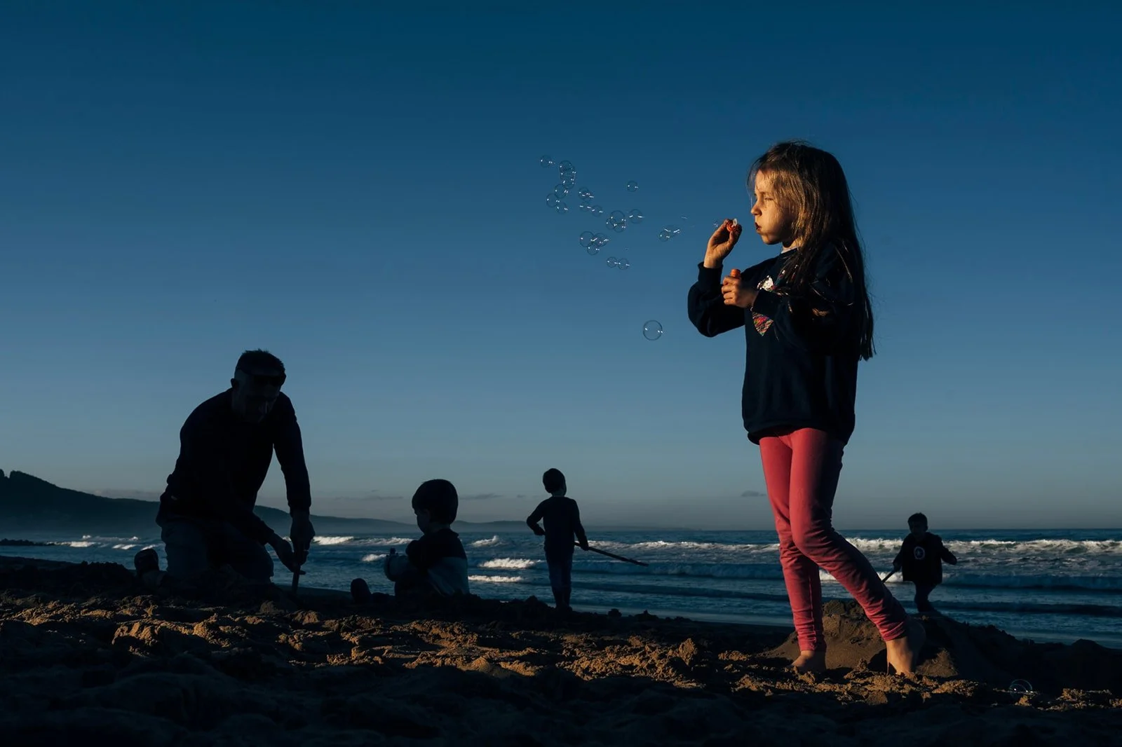 A girl standing on a beach blowing bubbles with the ocean and other children playing in the background during sunset.