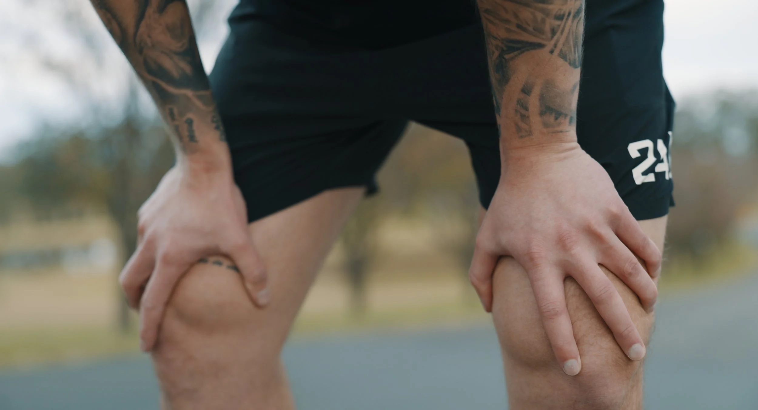 Person with tattoos leaning over with hands on knees, wearing black shorts, outdoors with blurred trees in background.