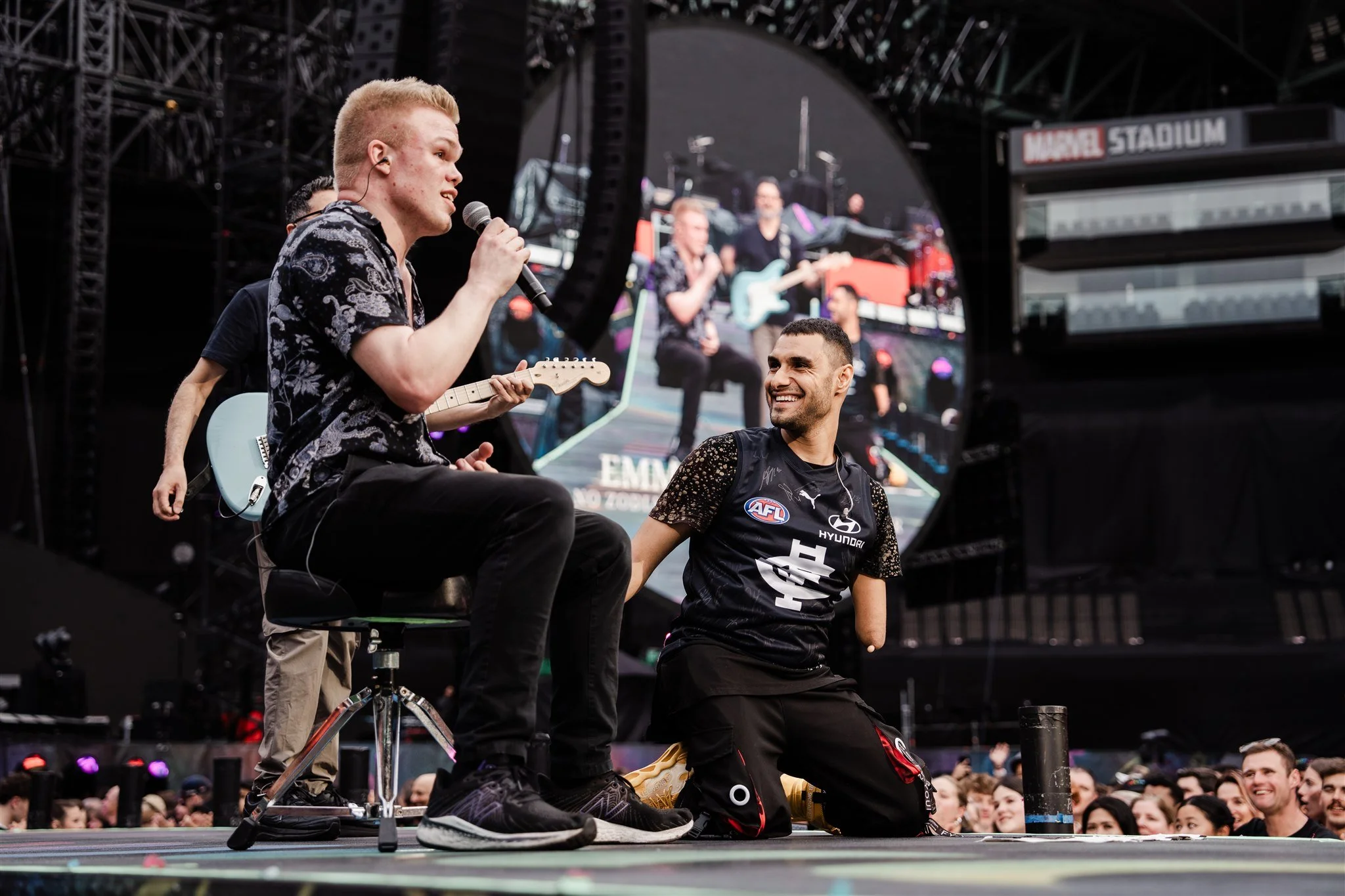 A man holding a microphone and sitting on a stool, smiling, dressed in a black sports jersey with an AFL logo, is on stage at a large outdoor event. Another man, also in sportswear, is kneeling in front of him, smiling and looking up. In the background, musicians are playing guitars and singing, with a large crowd watching.