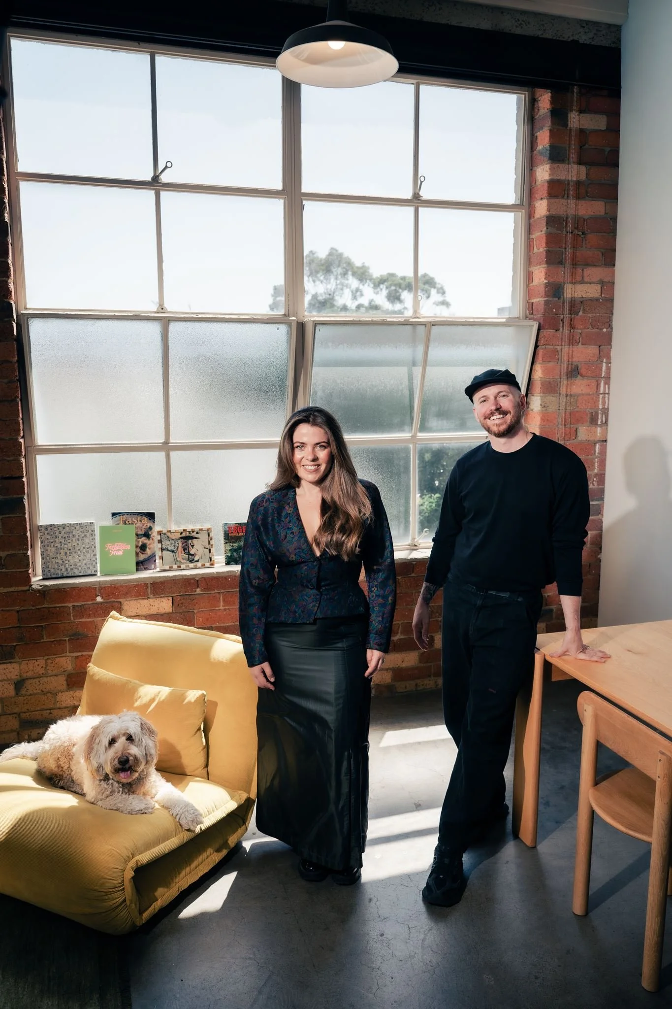A woman and a man standing inside a room with brick walls and large windows, a dog lying on a yellow couch in the foreground, and books and magazines on a ledge by the window.