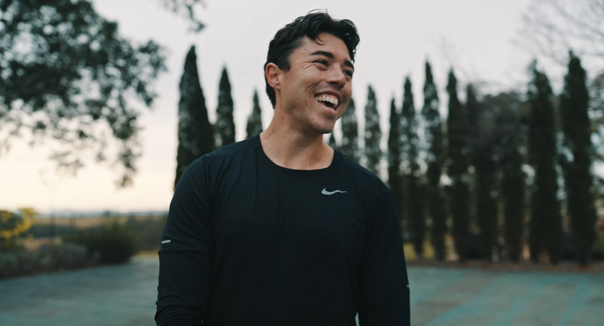 A man with dark hair, wearing a black Nike shirt, smiling outdoors with trees in the background.