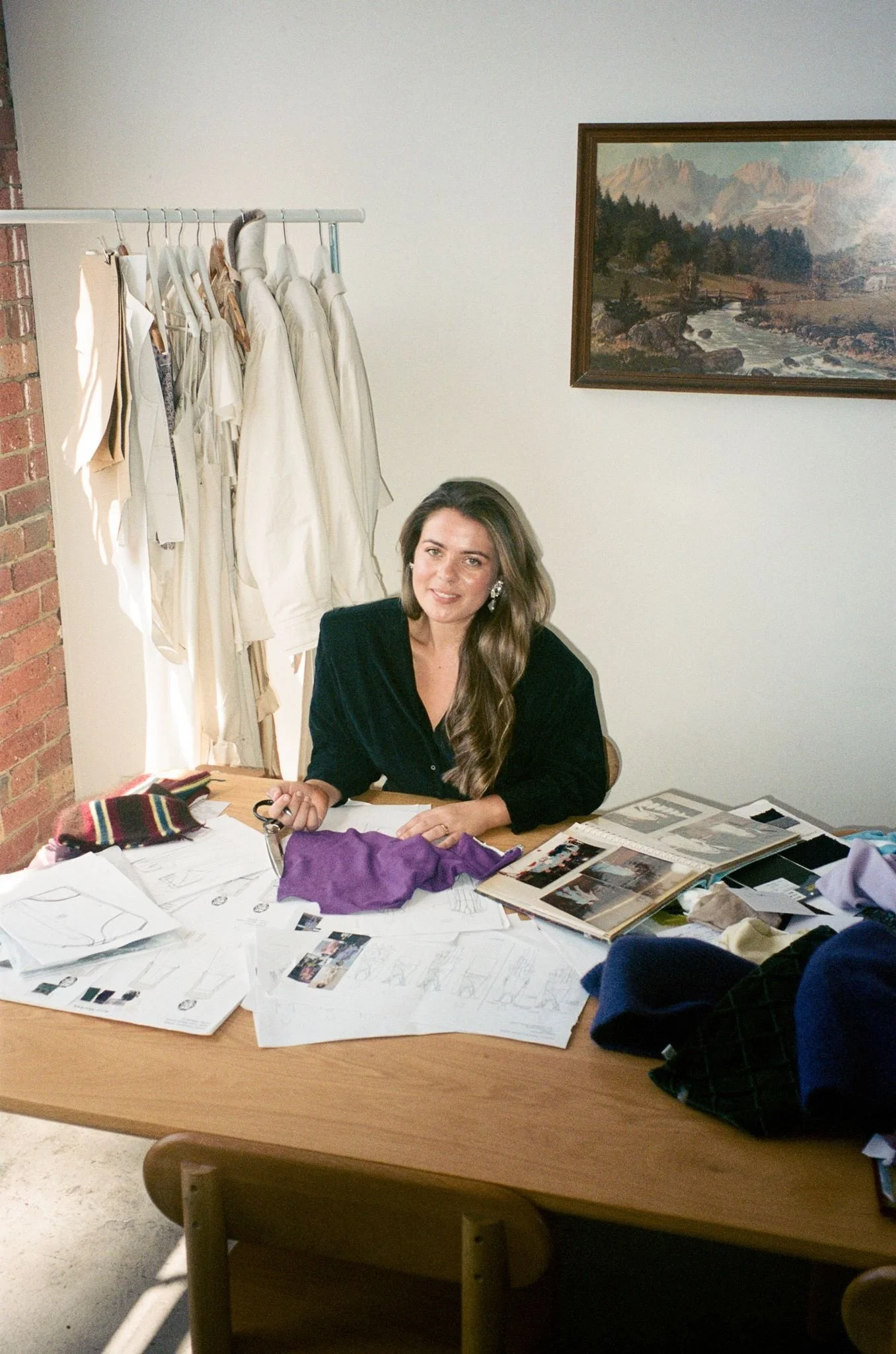 A woman with long, wavy brown hair, sitting at a cluttered table covered with papers, photographs, and fabric swatches, smiling and holding a pair of scissors. Behind her, there is a rack with white garments and a framed landscape painting of mountains, trees, river, and a bridge on a plain wall.