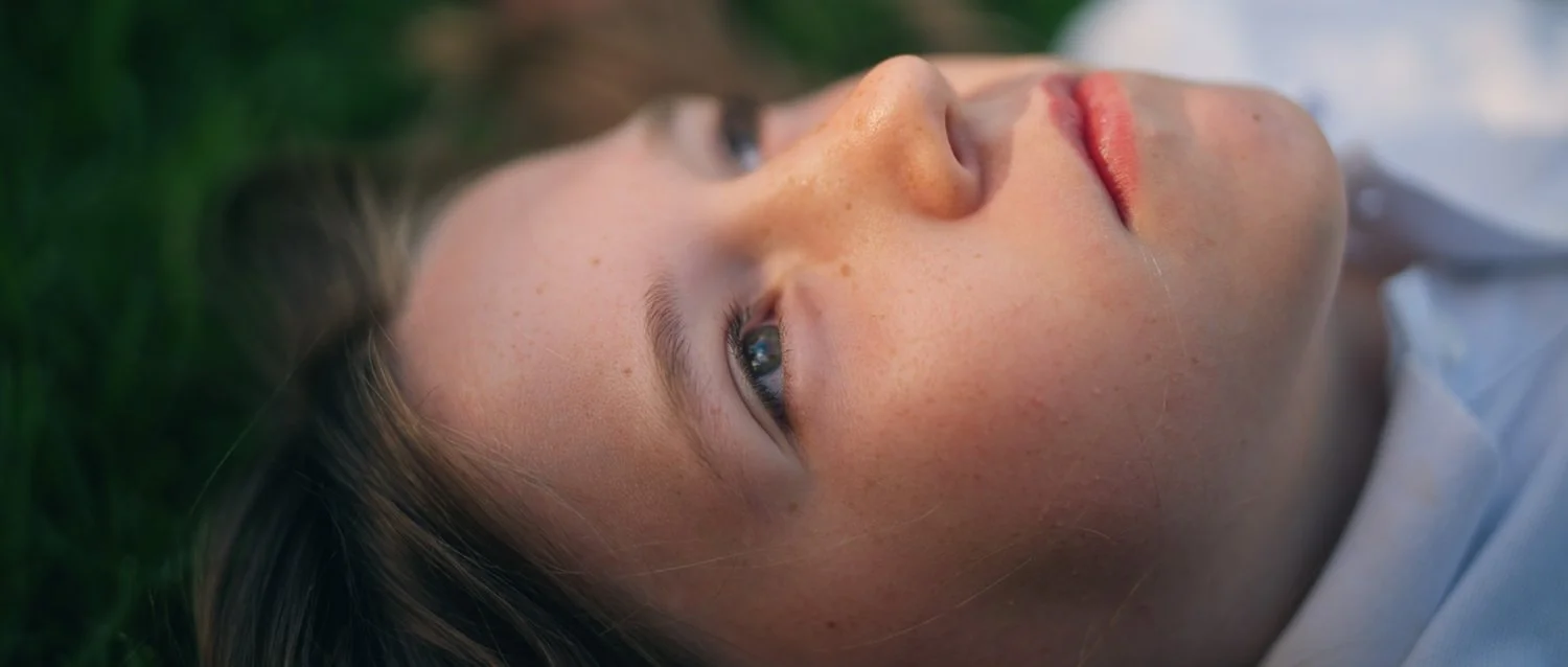 Film still from Things Will Be Different, showing a close-up of a young woman lying on the grass and looking upwards.