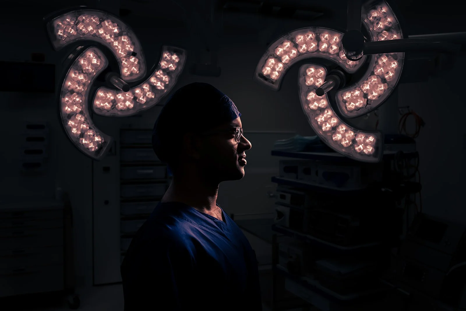 A healthcare professional wearing scrubs and glasses standing in a dark room with surgical lights overhead.