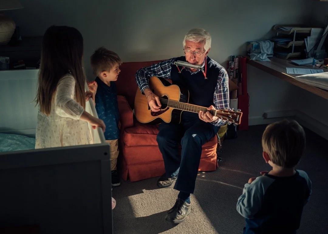 An older man playing an acoustic guitar and harmonica for three children in a dimly lit room.