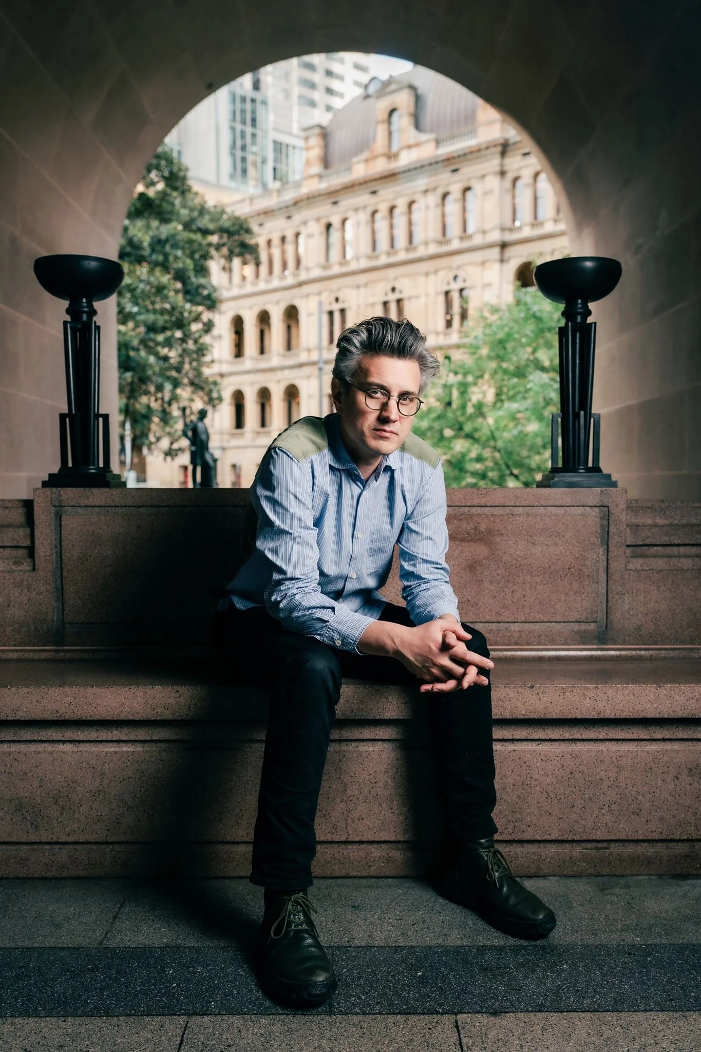 A man with glasses and gray hair sits on stone steps outside a historic building, with modern skyscrapers in the background. The scene is framed by an archway and flanked by two black lamps.