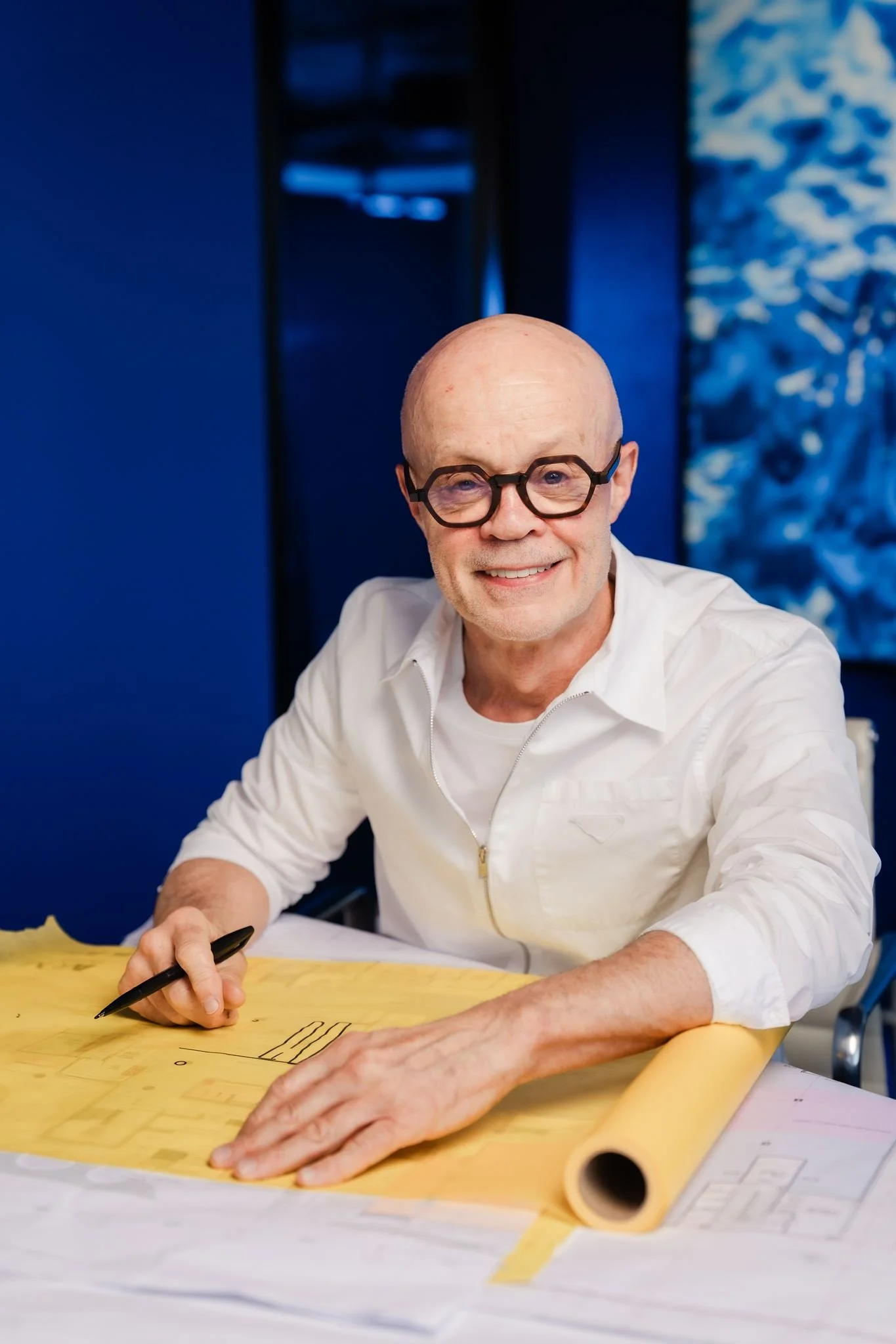 An older man with bald head and glasses sitting at a table, smiling, holding a pen, working on architectural plans or blueprints, with blue walls, a black structure, and a patterned blue and white wall in the background.