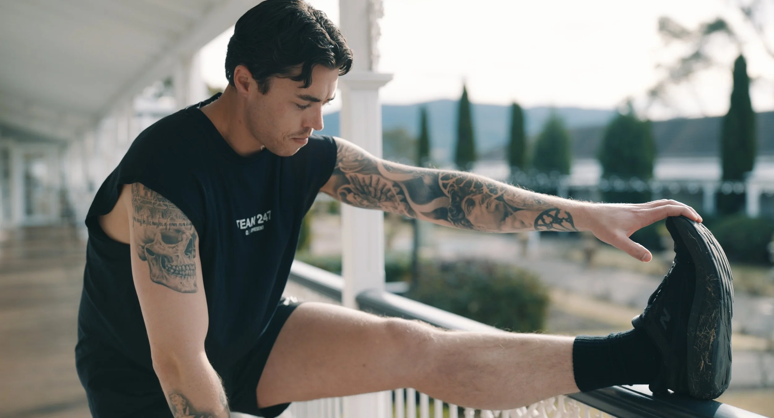 A young man with dark, wavy hair and tattoos on his arm, wearing a black sleeveless shirt and black shorts, sitting on a porch and tying his shoelace on his right shoe.