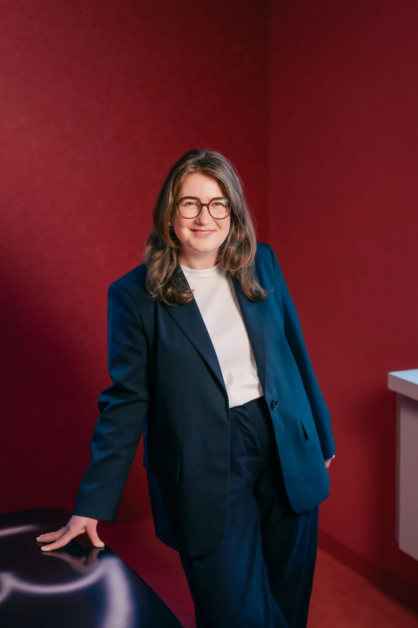 A woman with shoulder-length brown hair wearing glasses, a navy blazer, and navy trousers, leaning on a table with her left hand, standing in front of a red wall, smiling.