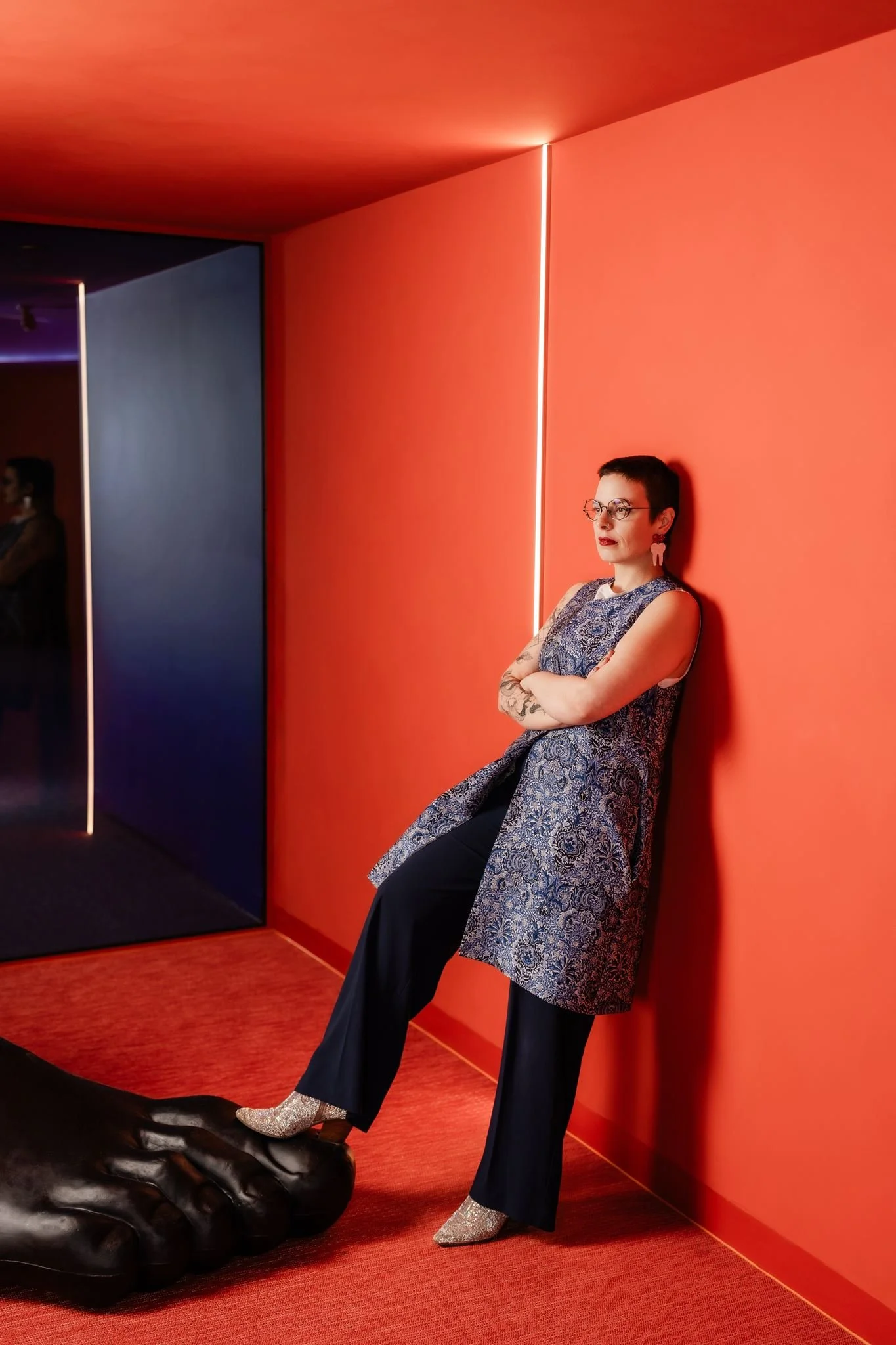 A woman with short dark hair and glasses sitting against a red wall with arms crossed, wearing a patterned sleeveless top and flared pants, with silver glitter shoes, and tattooed arms, in a modern interior with red carpet and neon lighting.