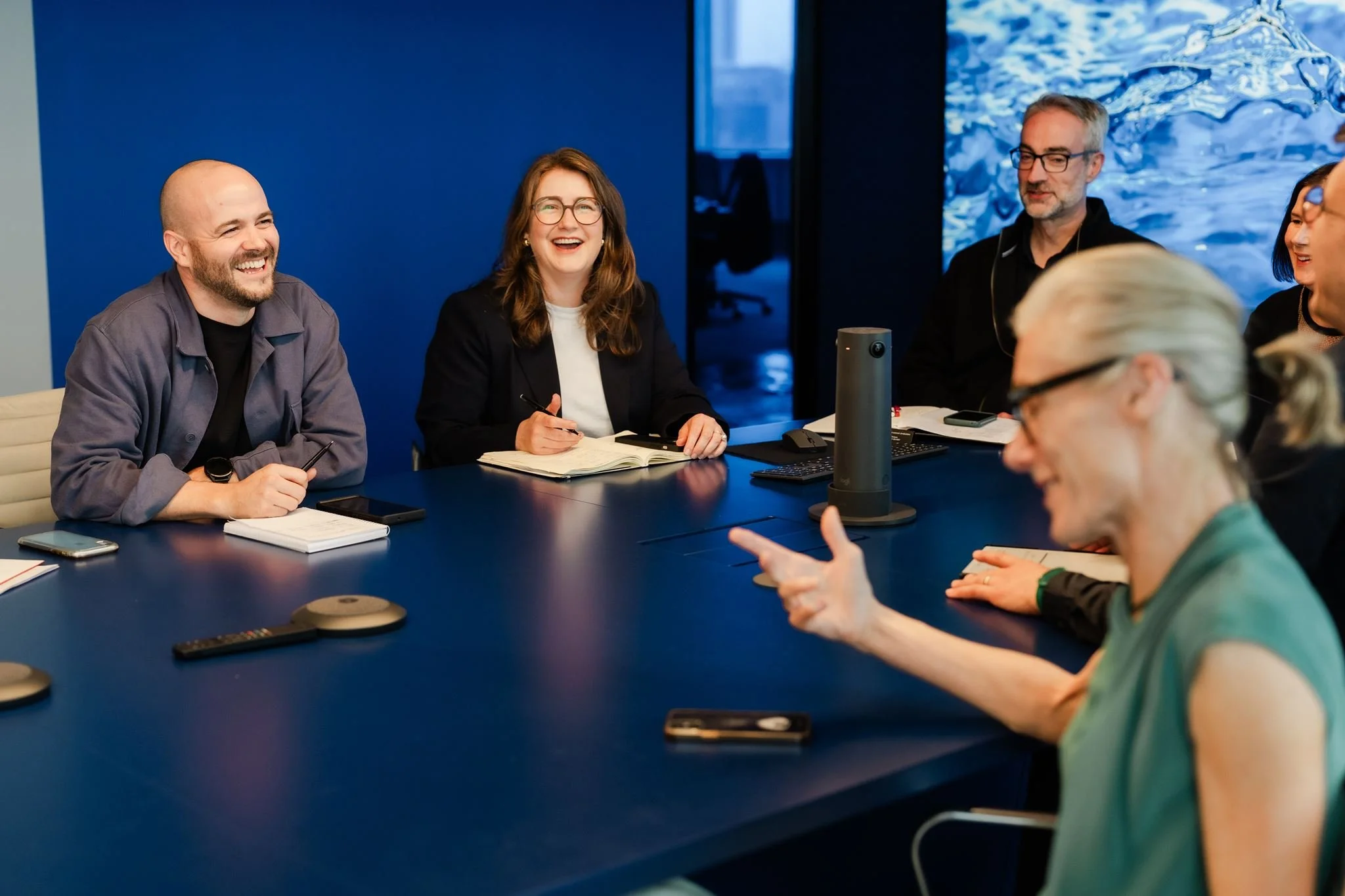 Group of diverse professionals in a meeting room, engaging in conversation, with a blue wall and a large digital screen in the background.