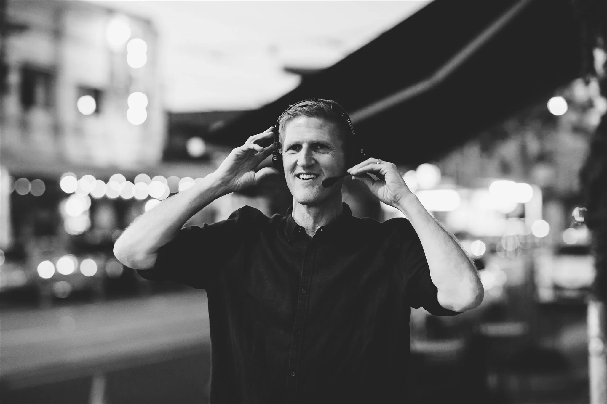 A black-and-white photo of a man wearing a headset in an urban setting at night, with blurred city lights in the background.