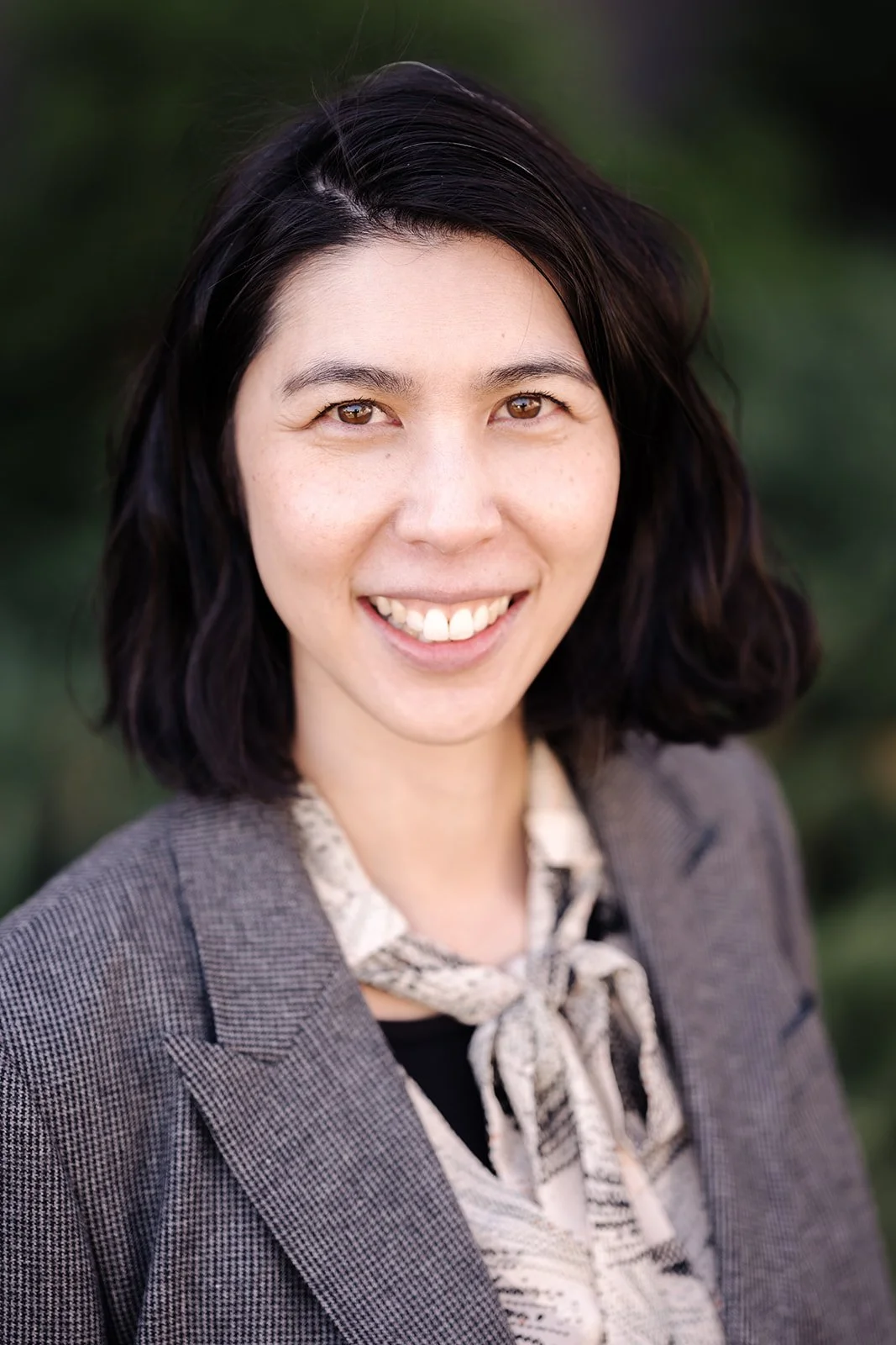 A smiling woman with shoulder-length dark hair and light skin, wearing a gray blazer and patterned scarf, standing outdoors with a blurred green background.