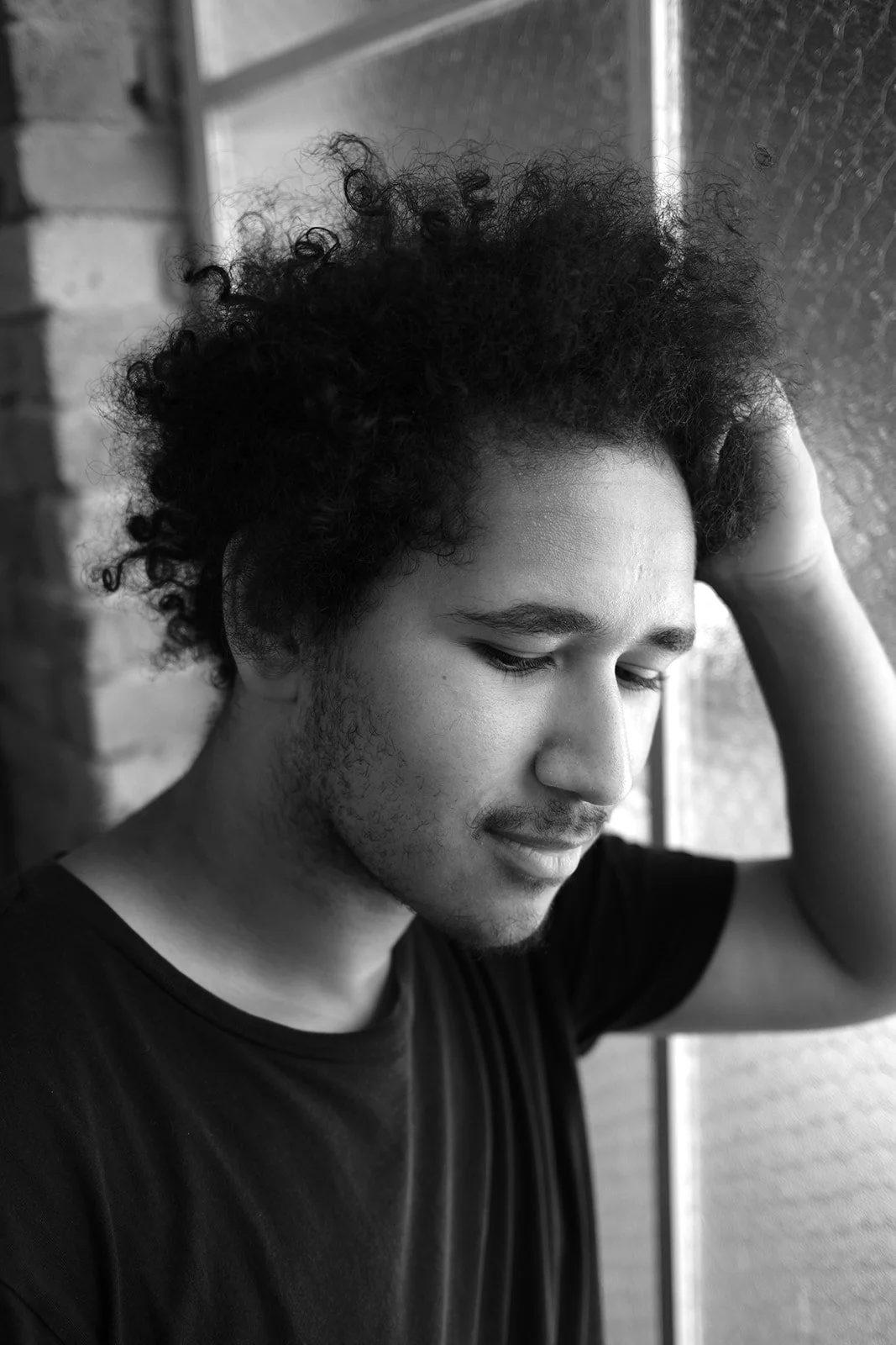 Black and white photo of a young man with curly hair, wearing a black t-shirt, with his eyes closed and one hand on his head, standing outdoors near a brick wall and a metal surface.
