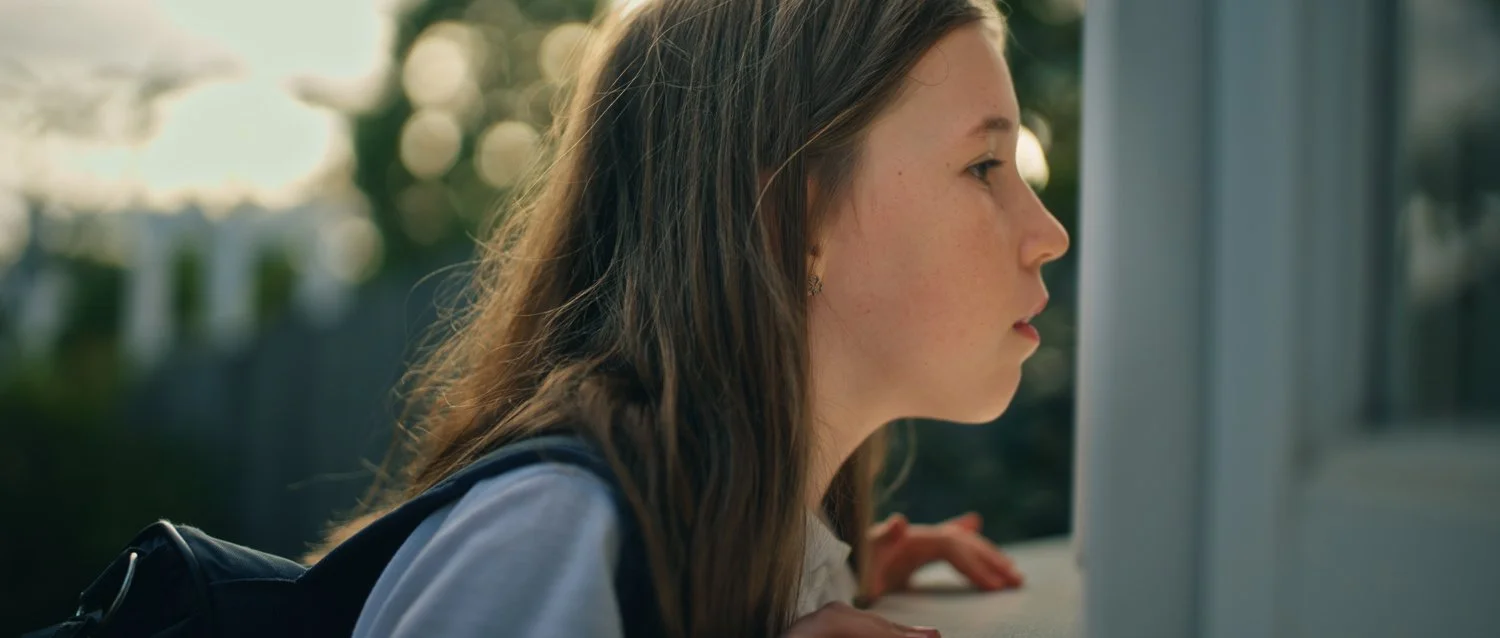Film still from Things Will Be Different, showing a young woman with long brown hair looking through a window during golden hour.