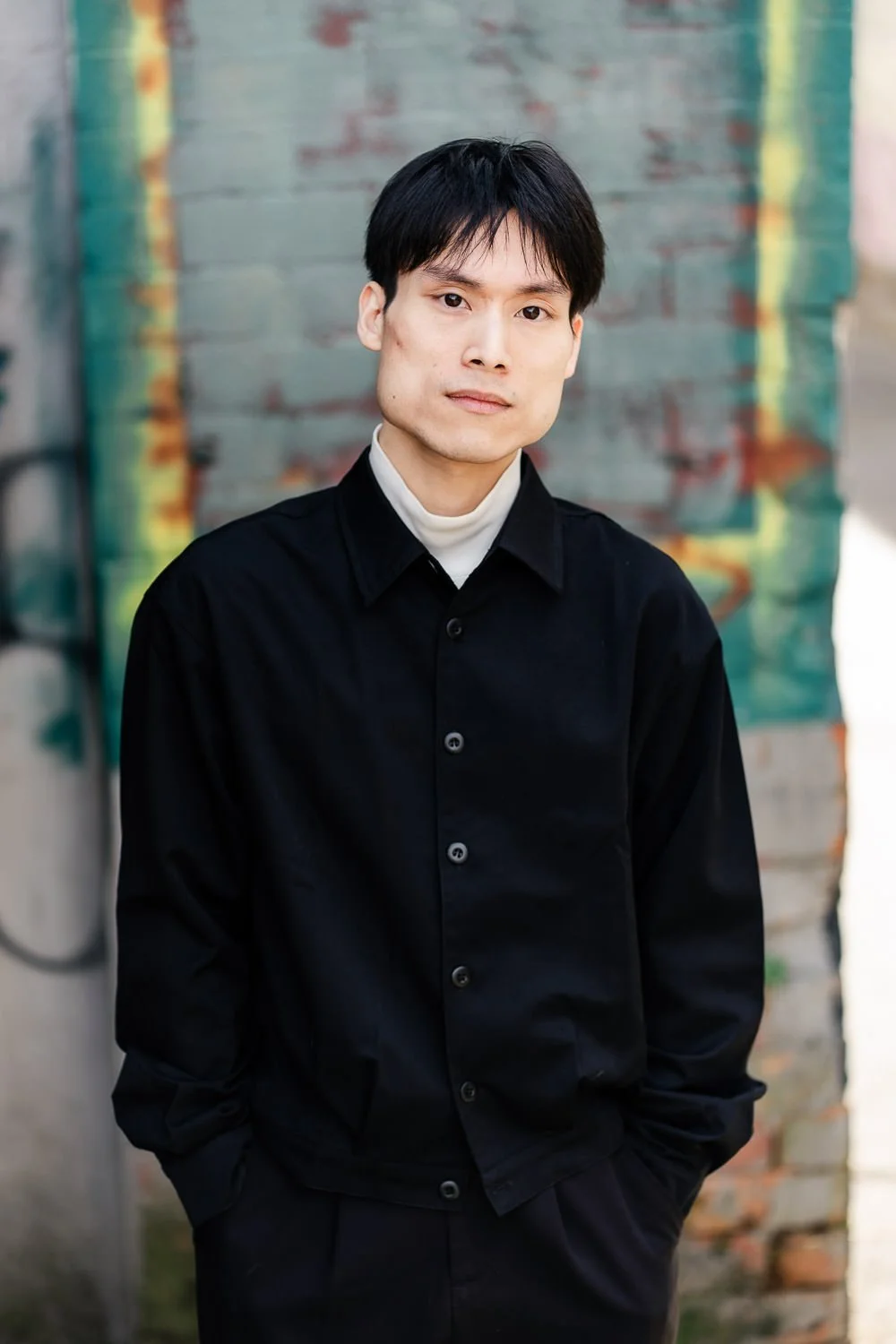 A young man with black hair wearing a black jacket and white turtleneck, standing against a graffiti-covered brick wall.