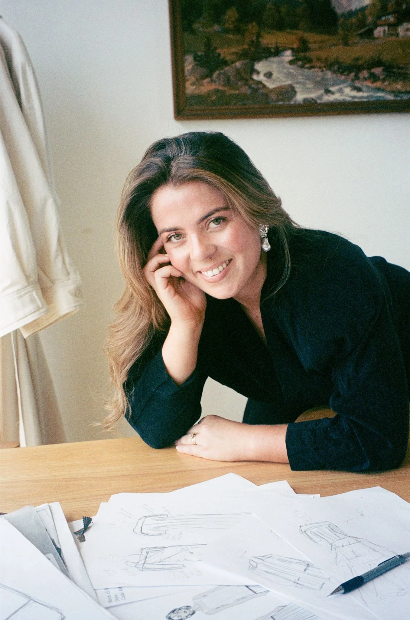 A woman with long wavy hair and earrings, wearing a dark shirt, sitting at a desk with sketches and papers, smiling at the camera with her head resting on her hand.