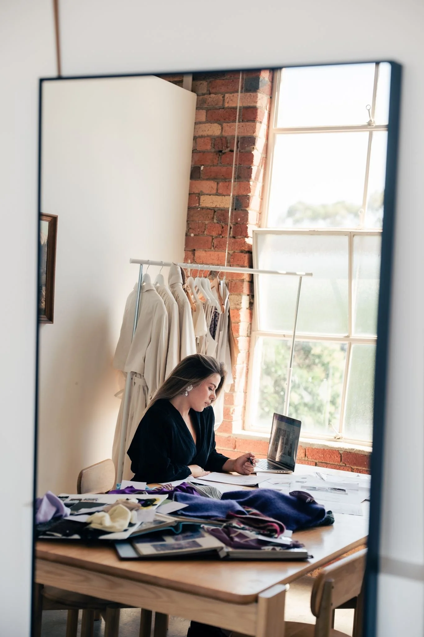 Portrait of Lauren Acciarito, founder and fashion designer of ACCI, working at her laptop in her studio, reflected in a mirror with garments hanging behind her.