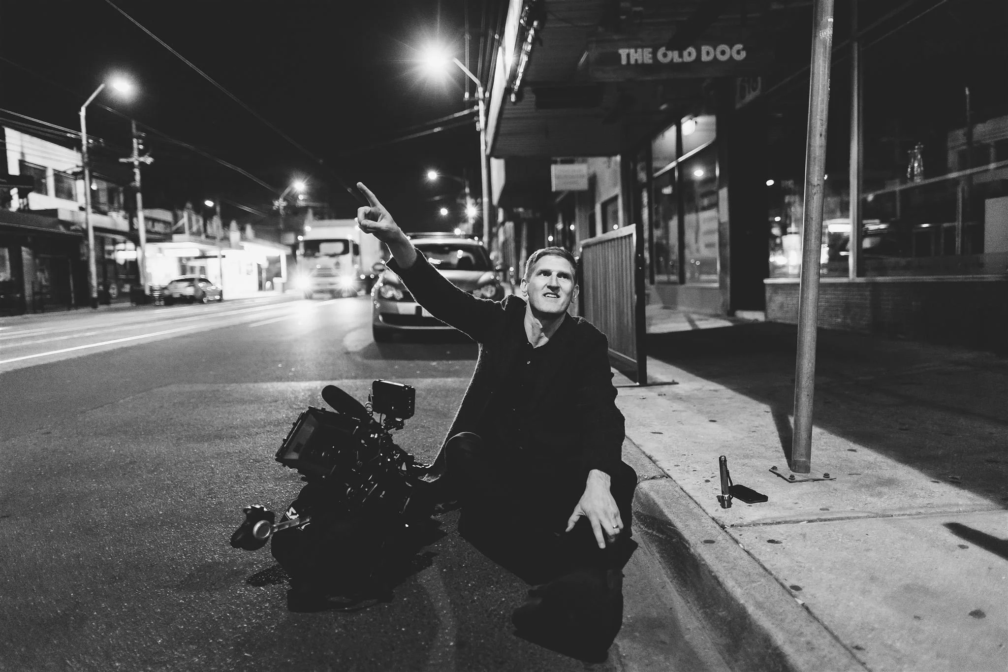 A man sitting on the sidewalk at night next to a film camera, gesturing upward with his right hand. He is in front of a storefront with a sign that reads 'The Old Dog.' Streetlights and parked cars are visible in the background.