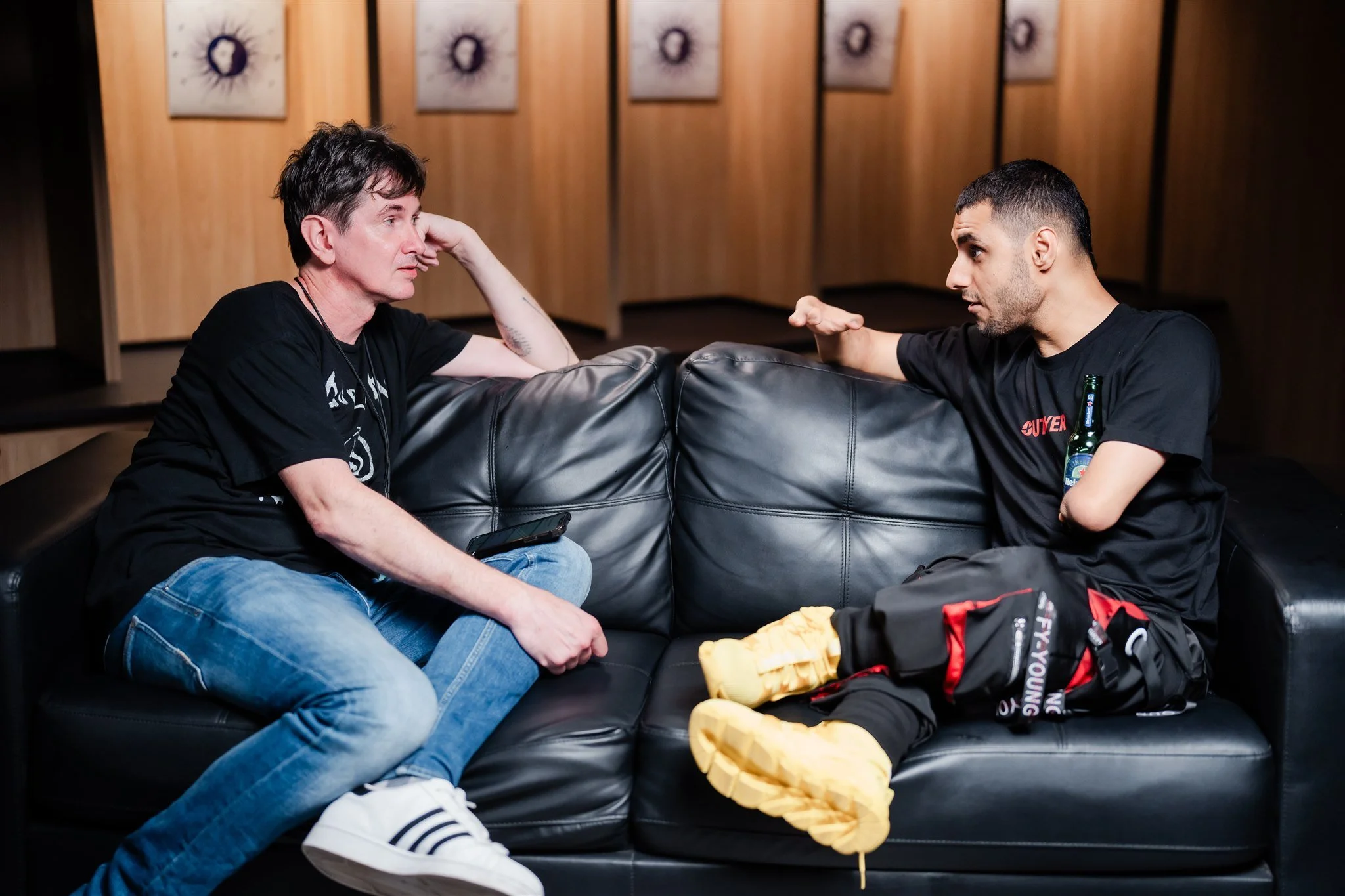 Emmanuel Kelly being interviewed on a black leather couch, seated in conversation with another man.