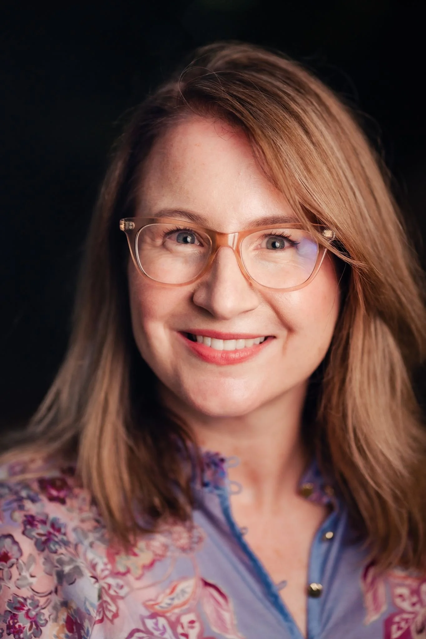 A woman with light brown hair and glasses, smiling, wearing a colorful floral top, against a black background.