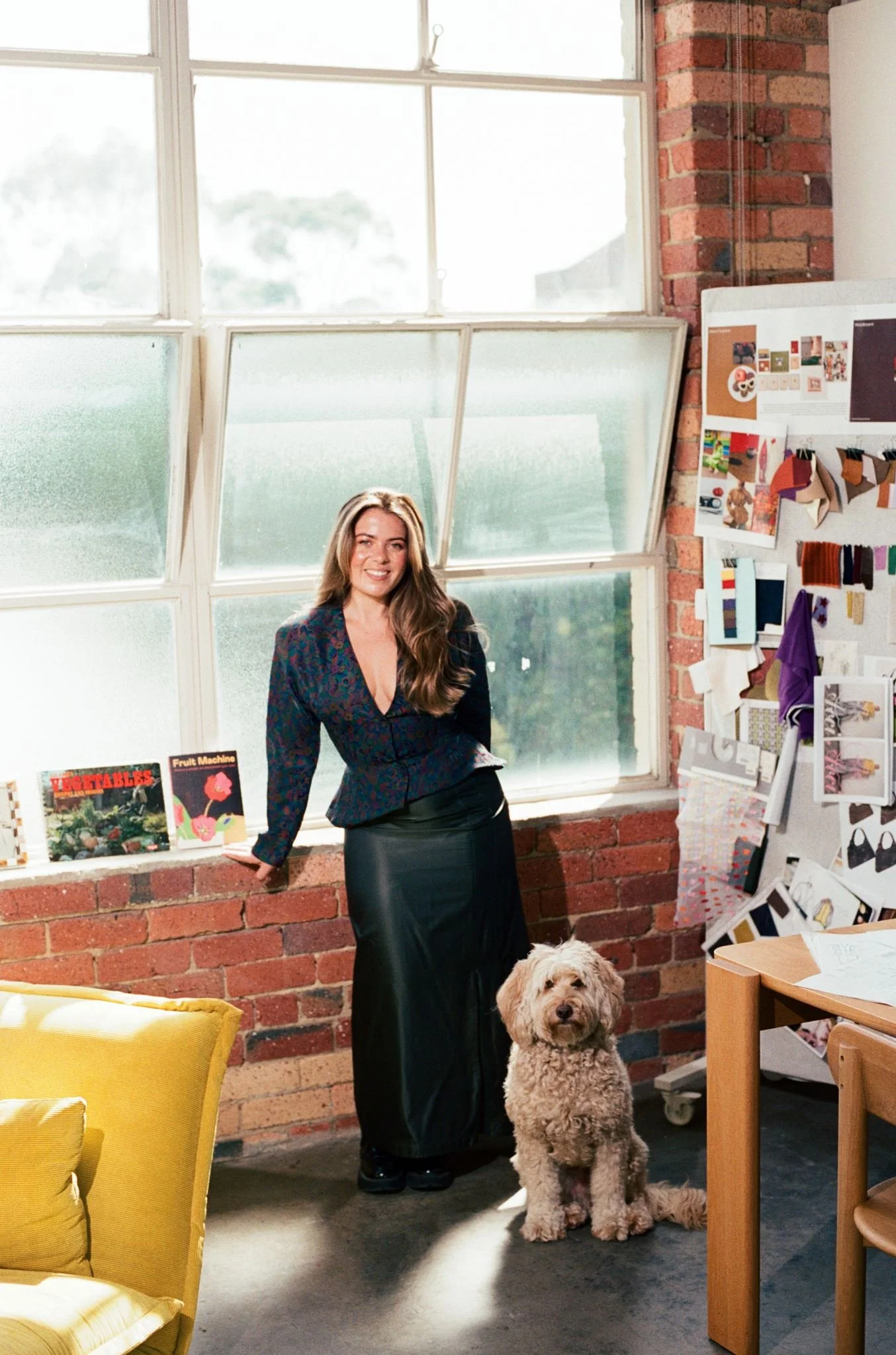 A woman with long, wavy hair wearing a navy blazer and black skirt standing inside a room with brick walls and large window panes. She is smiling at the camera with her hand resting on a brick ledge. A curly-haired dog with light brown fur sits in front of her, looking directly at the camera. There is an art display on the wall to her right and a yellow armchair visible at the bottom left corner.