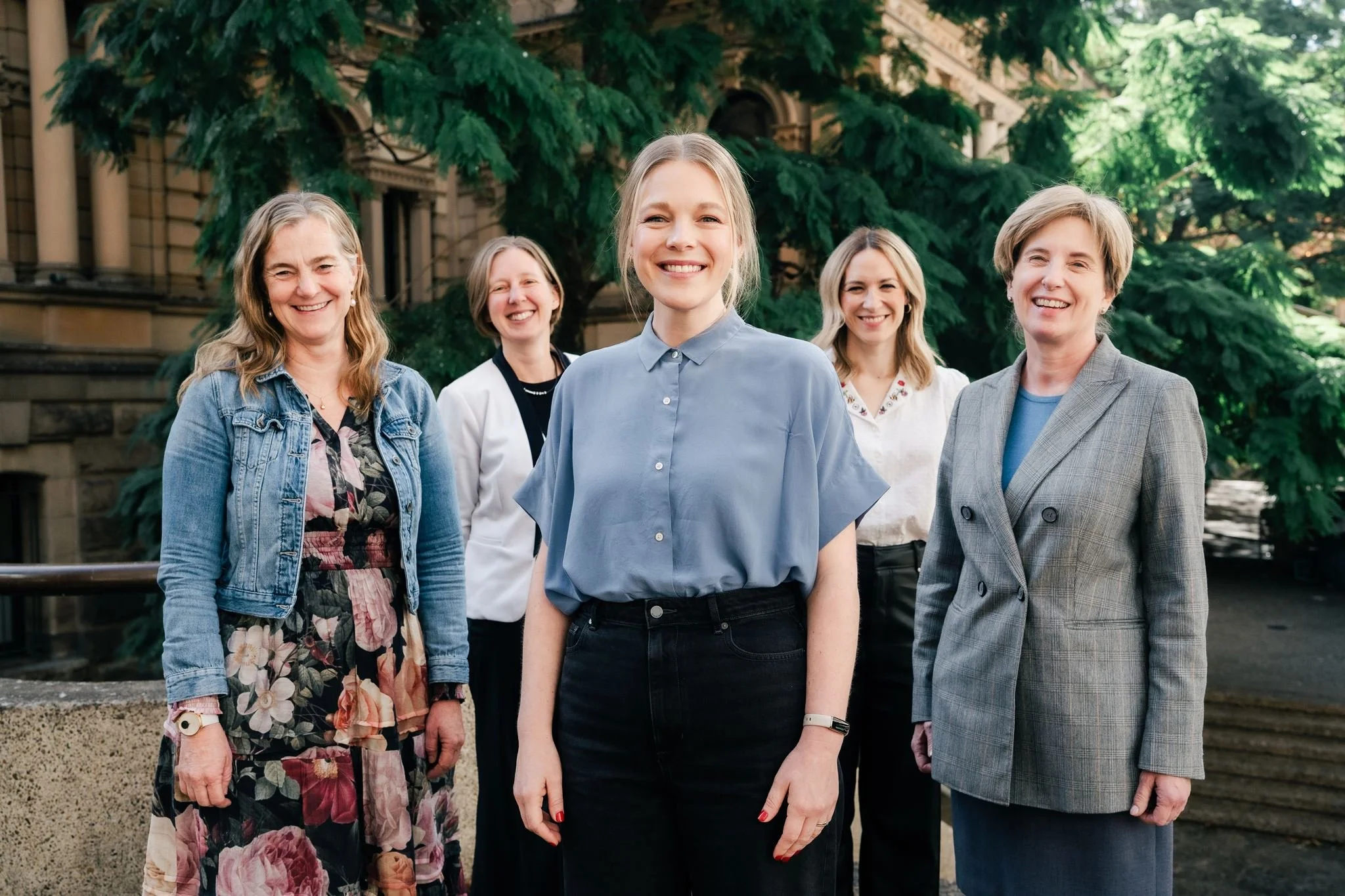 Group of five women standing outdoors in front of trees and historic buildings, smiling at the camera.