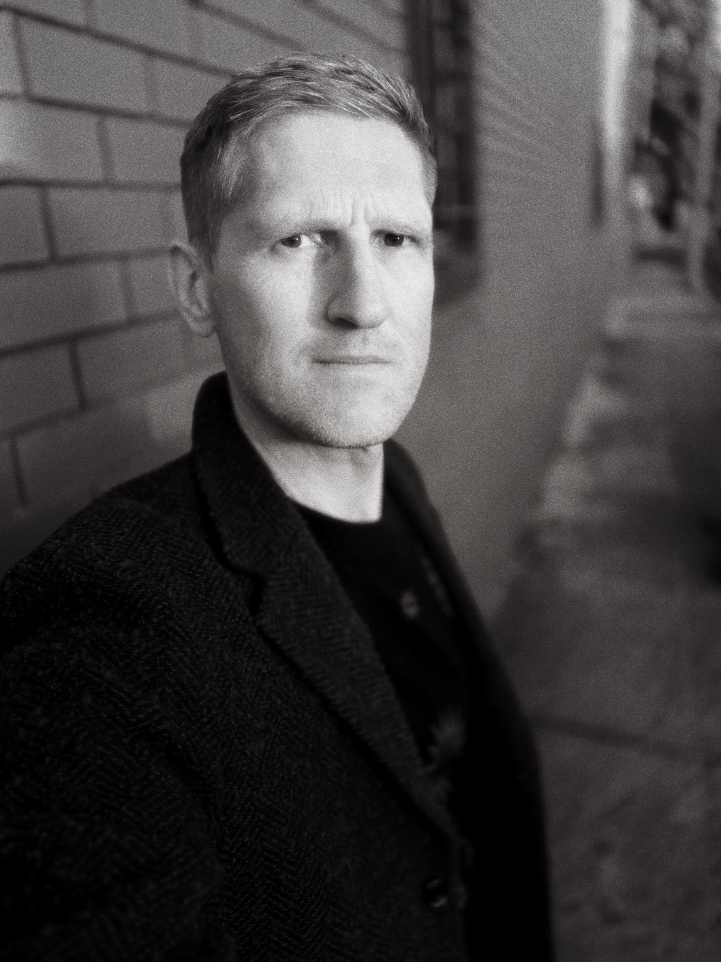 Black-and-white portrait of Melbourne director, cinematographer and photographer Jerome Cole, standing against a brick wall outdoors.