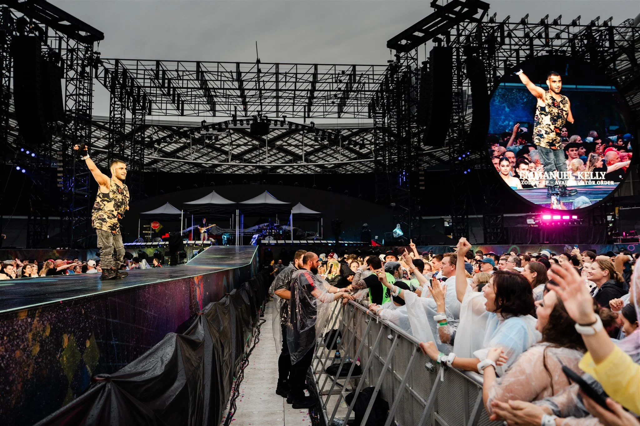 Emmanuel Kelly performing on stage in front of a crowd, with a large screen behind him displaying his name.