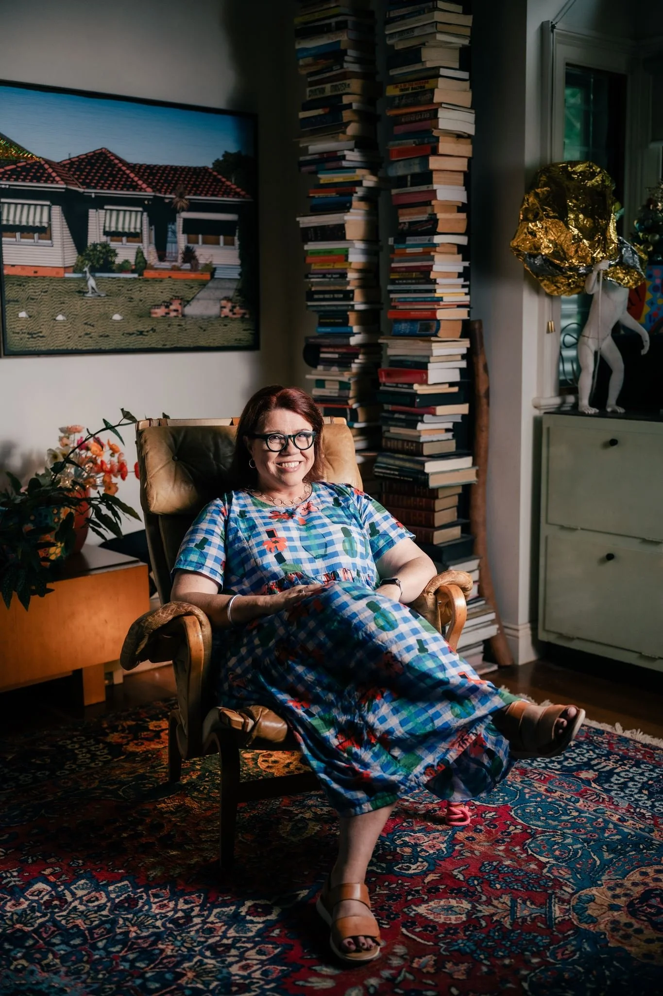 A smiling woman with glasses sitting in a cozy, decorated room with a large bookshelf, a painting of a house, a floral chair, and a colorful rug.