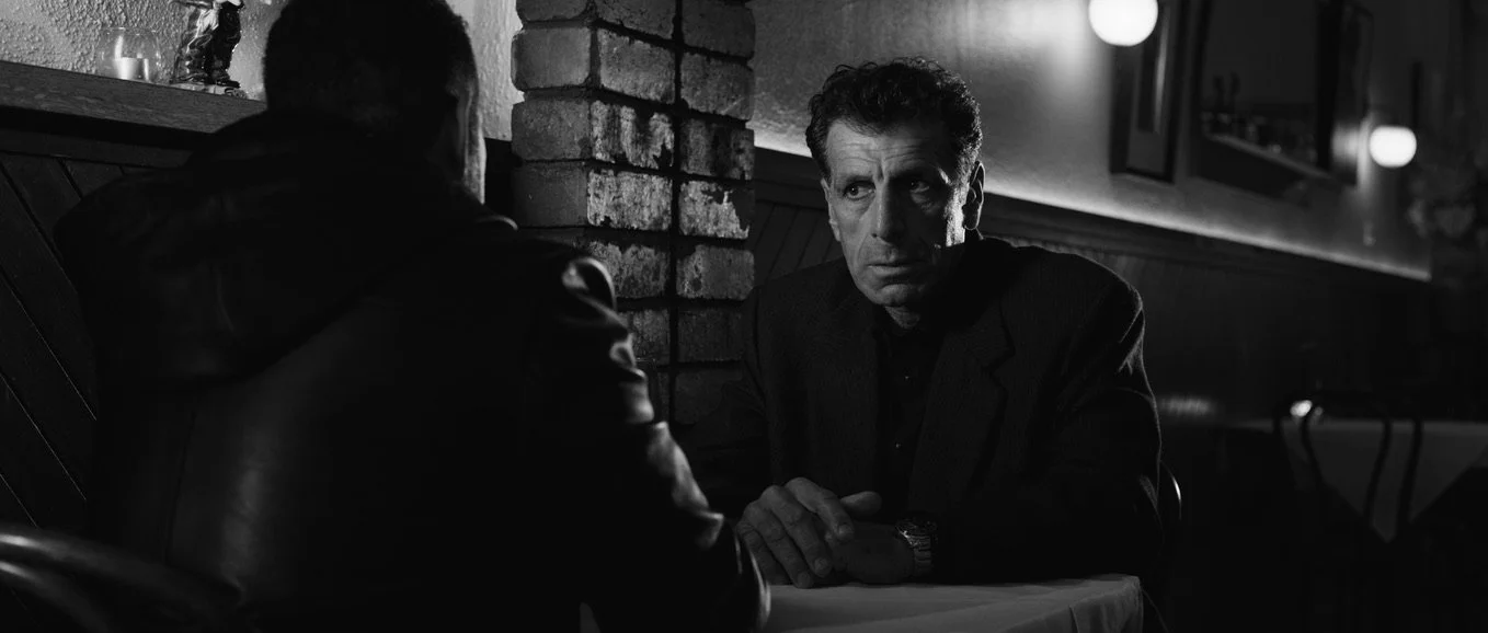 Film still from Cugini, showing two men seated across from each other at a table in a dimly lit restaurant.