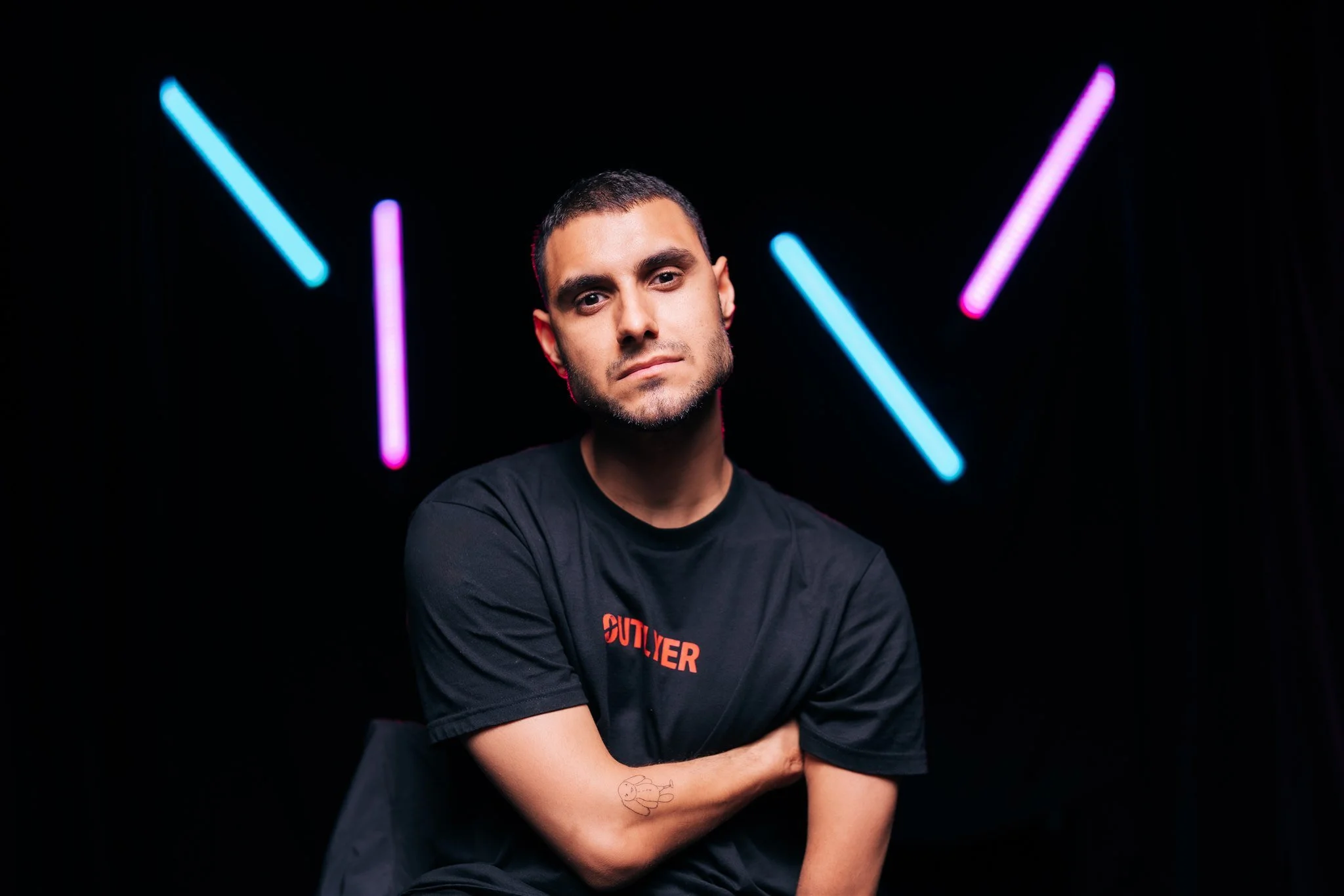 Landscape portrait of Emmanuel Kelly, standing with arms crossed against a black background with blue and pink neon lights.