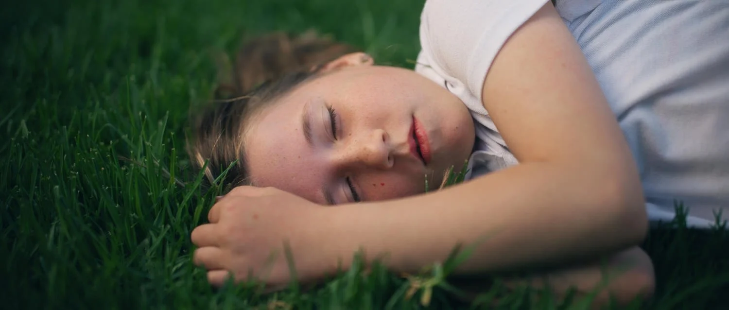 Film still from Things Will Be Different, showing a young girl with freckles asleep on the grass with her head resting on her arm.