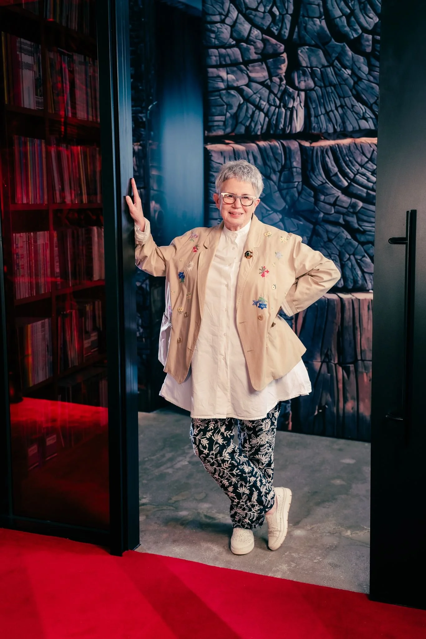 An older woman with short gray hair and glasses smiling, standing next to a dark wall with a large abstract blue and black textured painting or mural behind her. She is wearing a beige jacket with colorful embroidery, a white shirt, patterned black and white pants, and white sneakers.