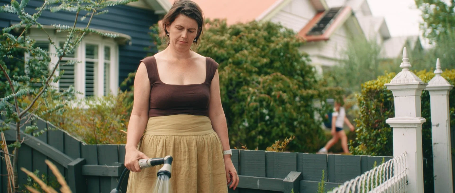 Film still from *Things Will Be Different*, showing a woman watering plants outside a blue house as a girl walks in the background.