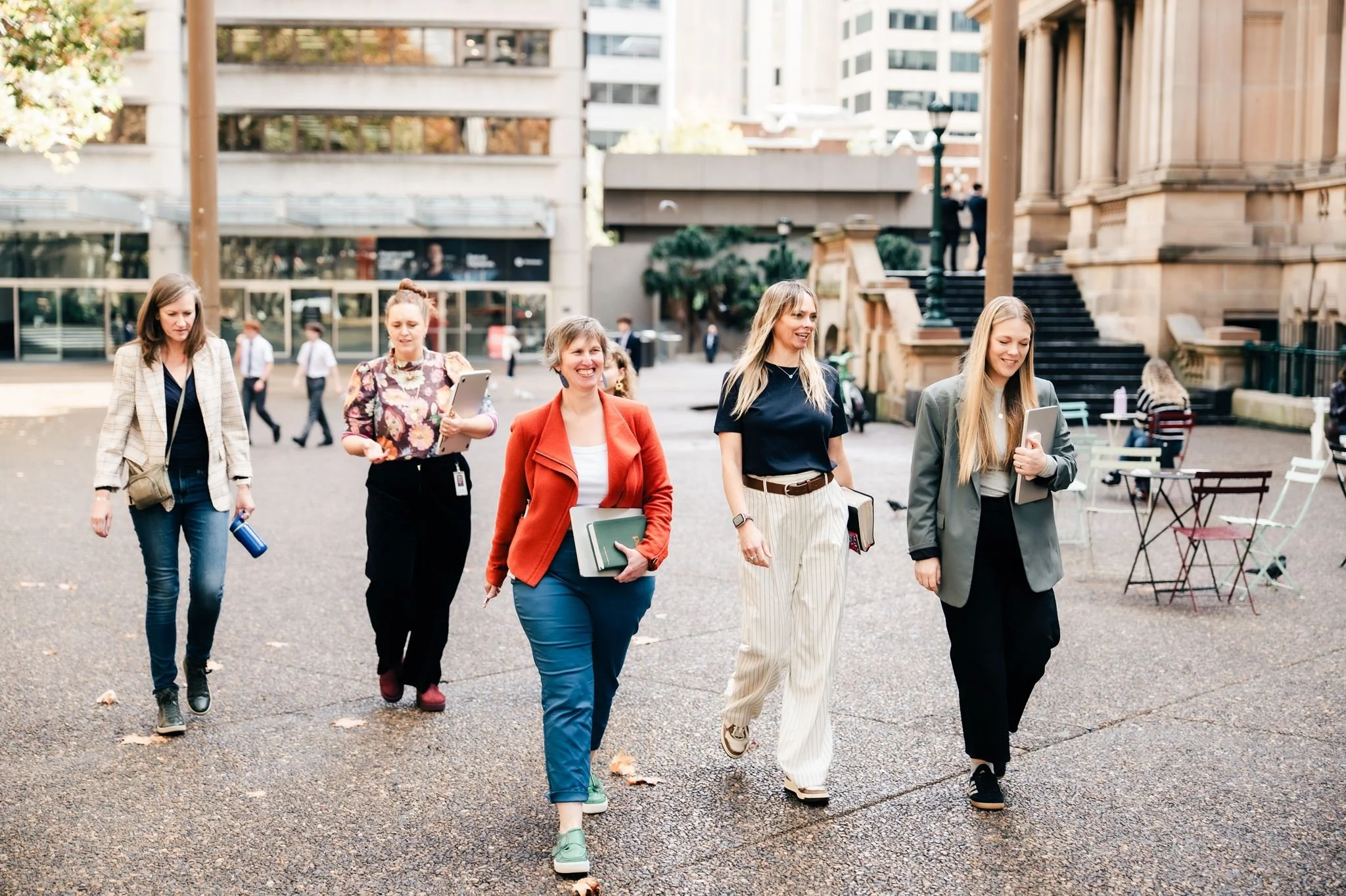 Group of women walking outdoors in an urban setting with buildings and stairs in background.