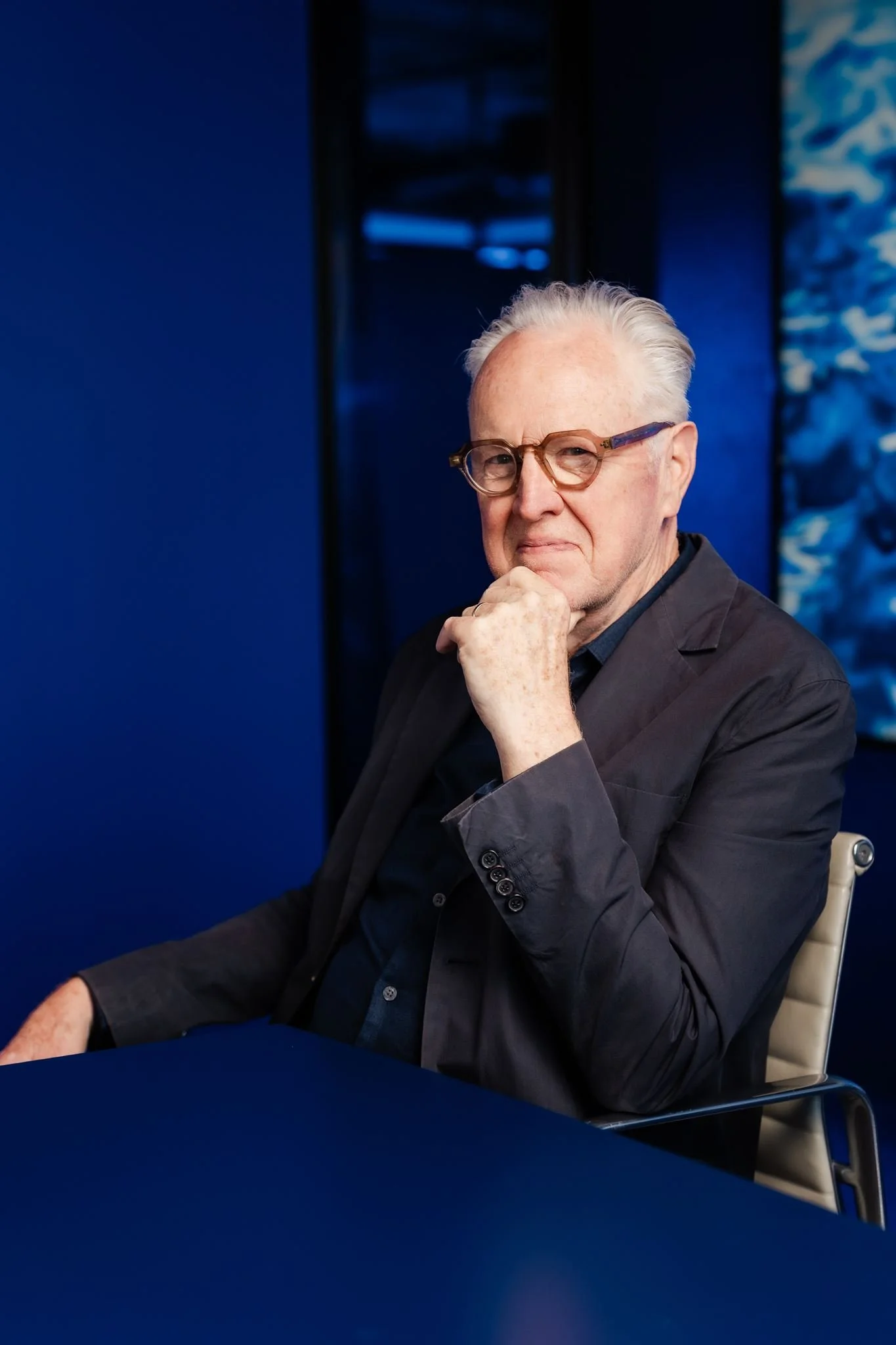Portrait of Howard Raggatt, co-founder of ARM Architecture, seated at a blue table in a blue-toned interior.