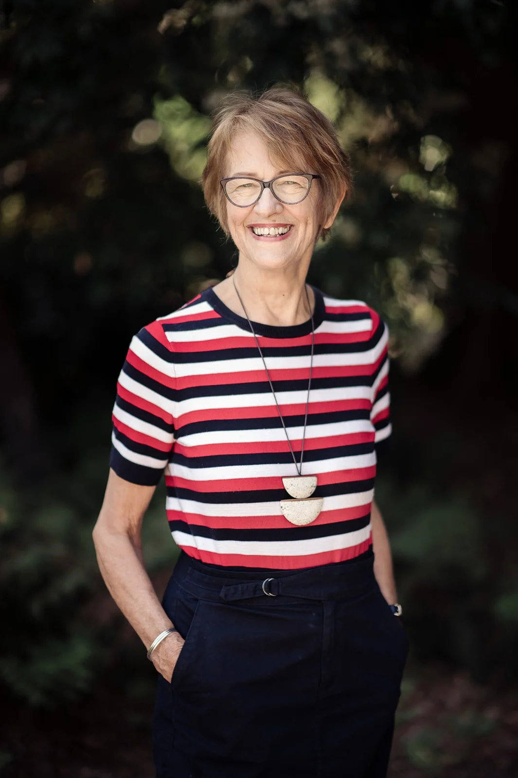 An older woman with short, light brown hair and glasses, smiling, wearing a red, white, and black striped shirt, dark pants, and a long necklace, outdoors with a blurred green background.