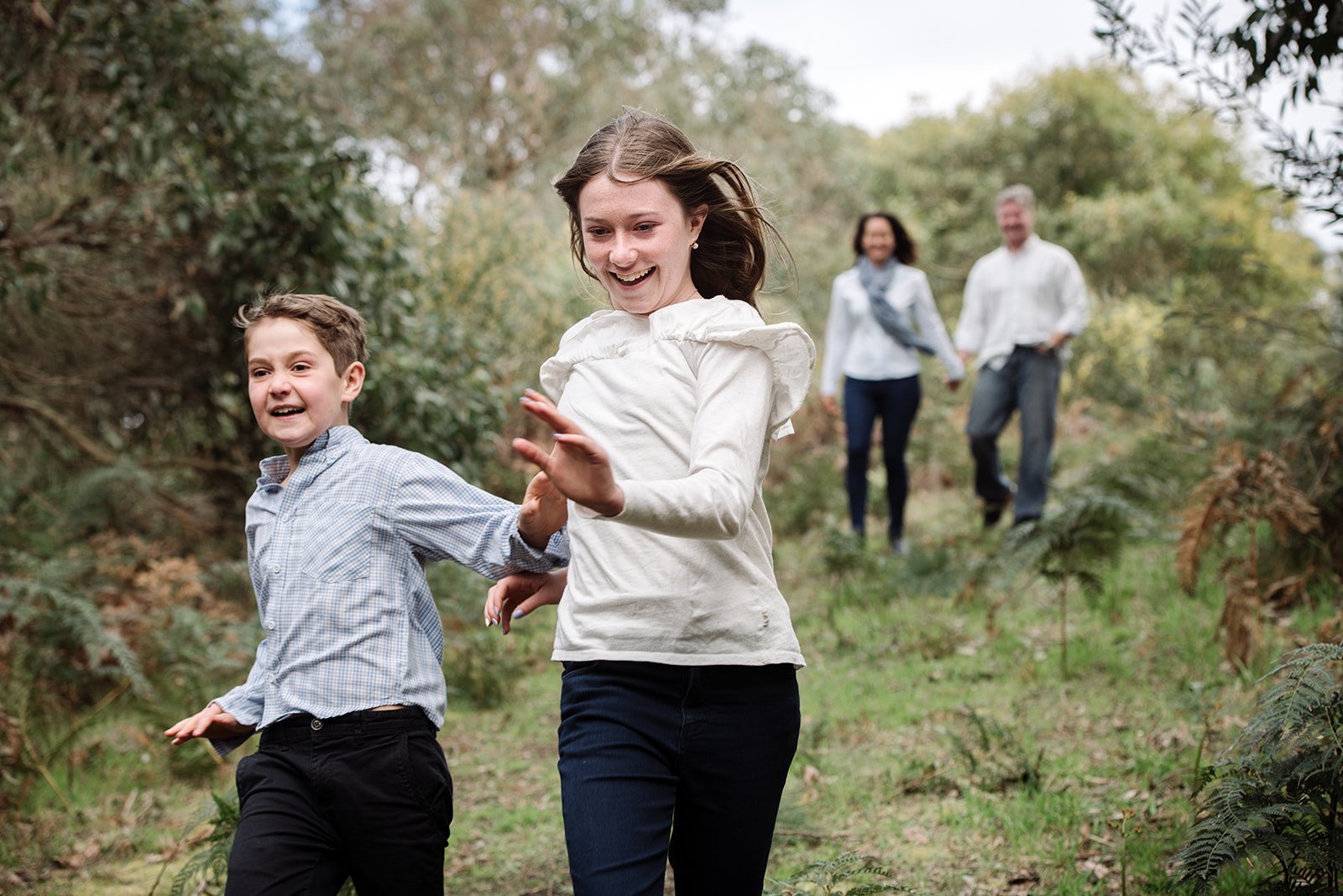 Four people, two children and two adults, are enjoying a walk outdoors in a forested area, with the children running ahead while smiling and the adults walking behind, hand in hand.
