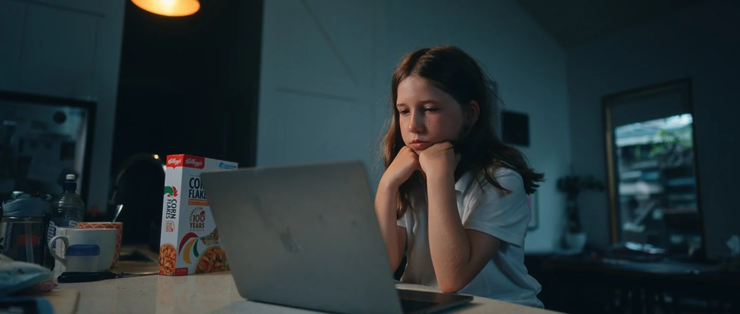 Film still from Things Will Be Different, showing a young girl sitting at a kitchen counter, looking at a laptop with a contemplative expression.