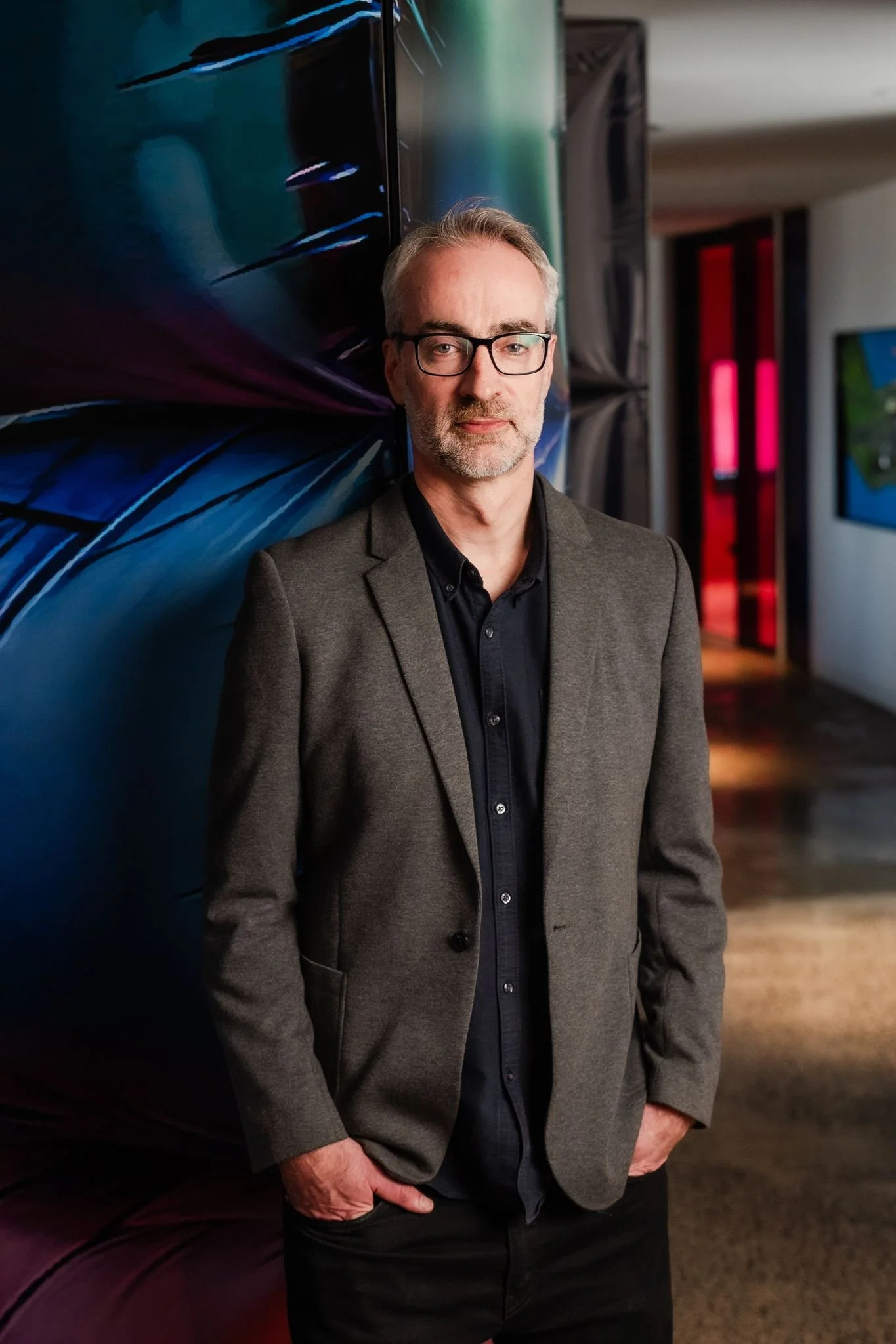 A middle-aged man with gray hair, beard, and glasses wearing a gray blazer over a black shirt, standing indoors against a colorful abstract background.