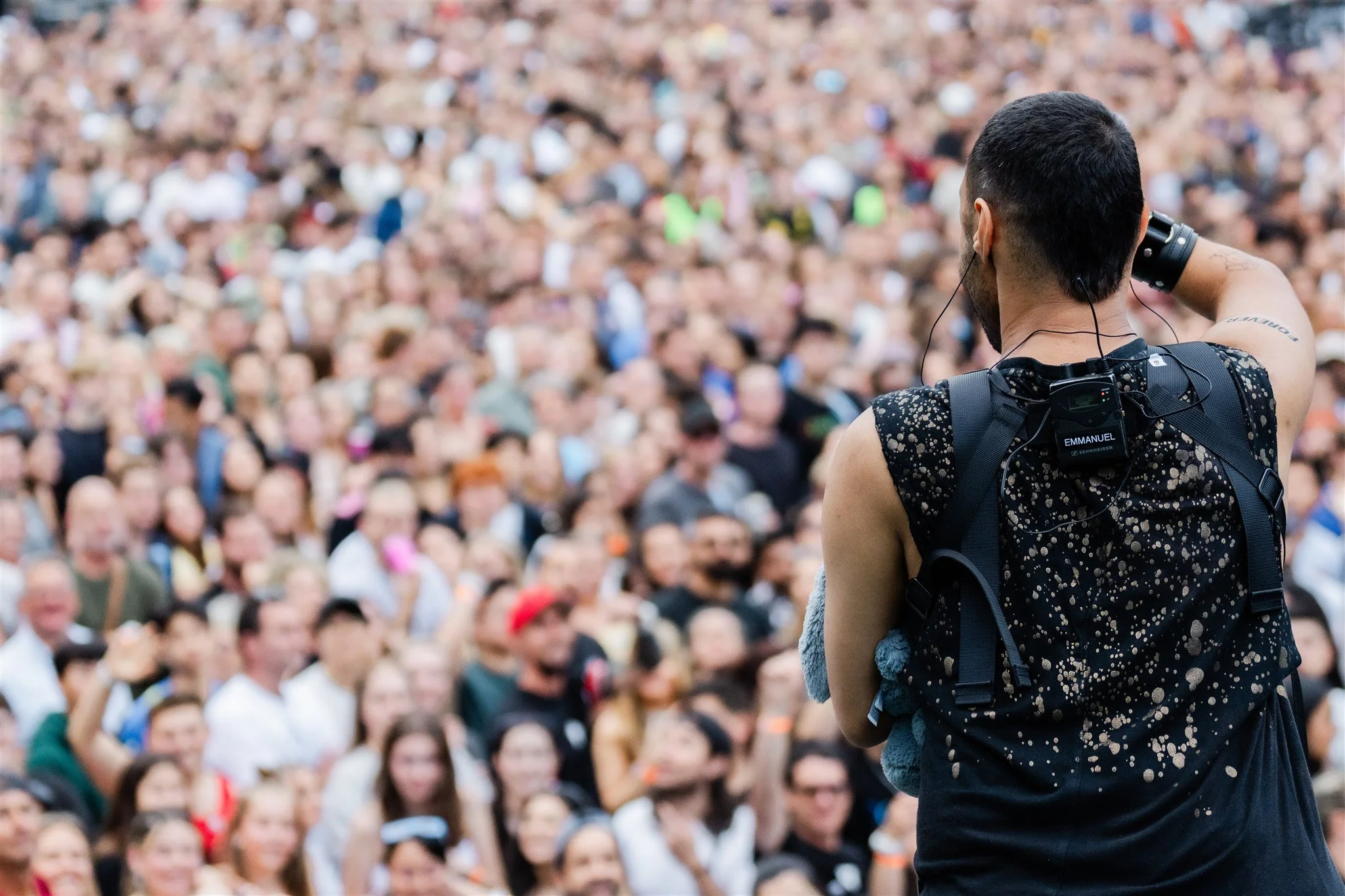 A person with short dark hair, wearing a black sleeveless shirt with a speckled design, is facing a large crowd of people in an outdoor setting. The individual has a backpack and earphones, with the back of their head and right arm visible.