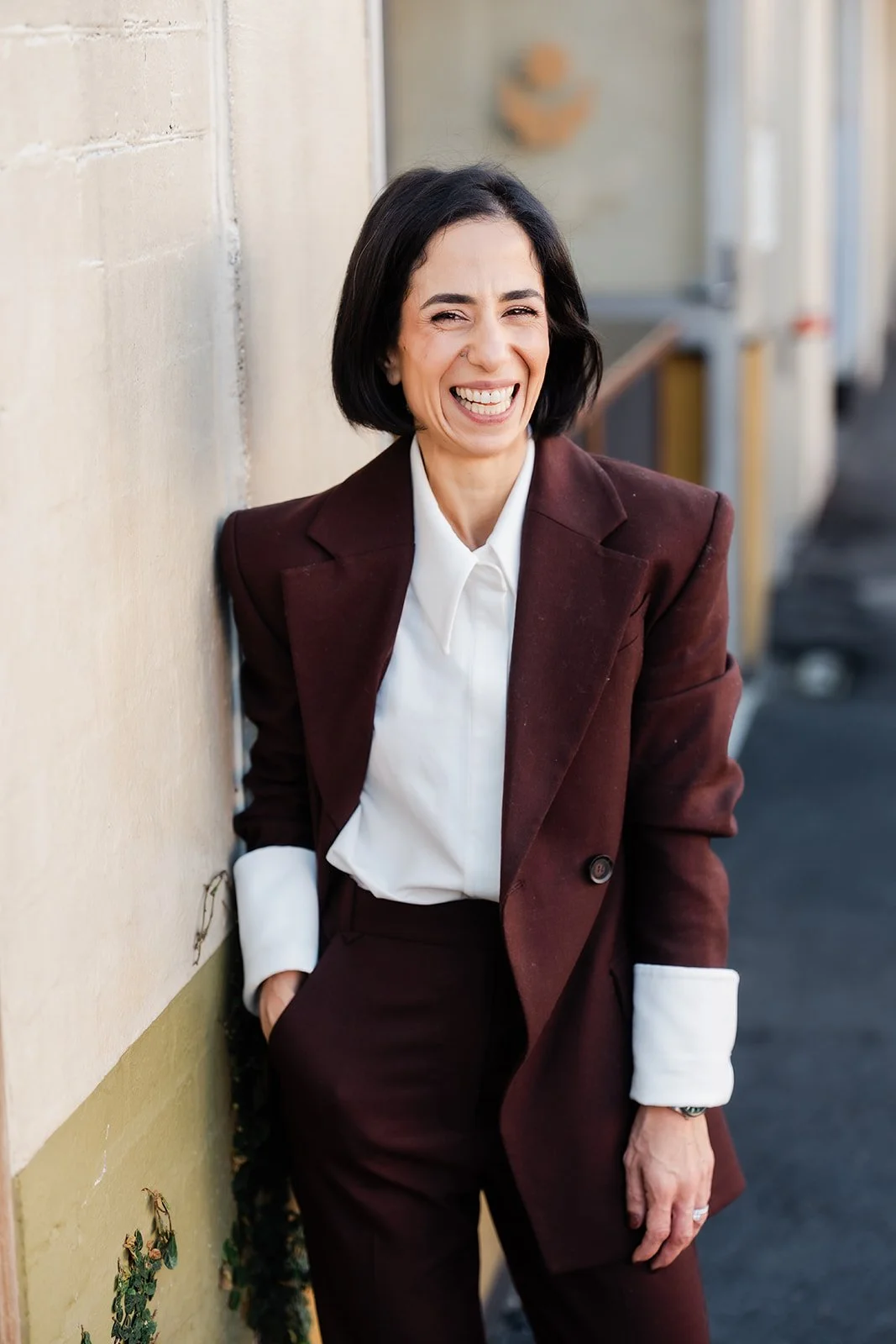 Smiling woman with short black hair in a brown suit with white shirt, leaning against a wall outdoors.