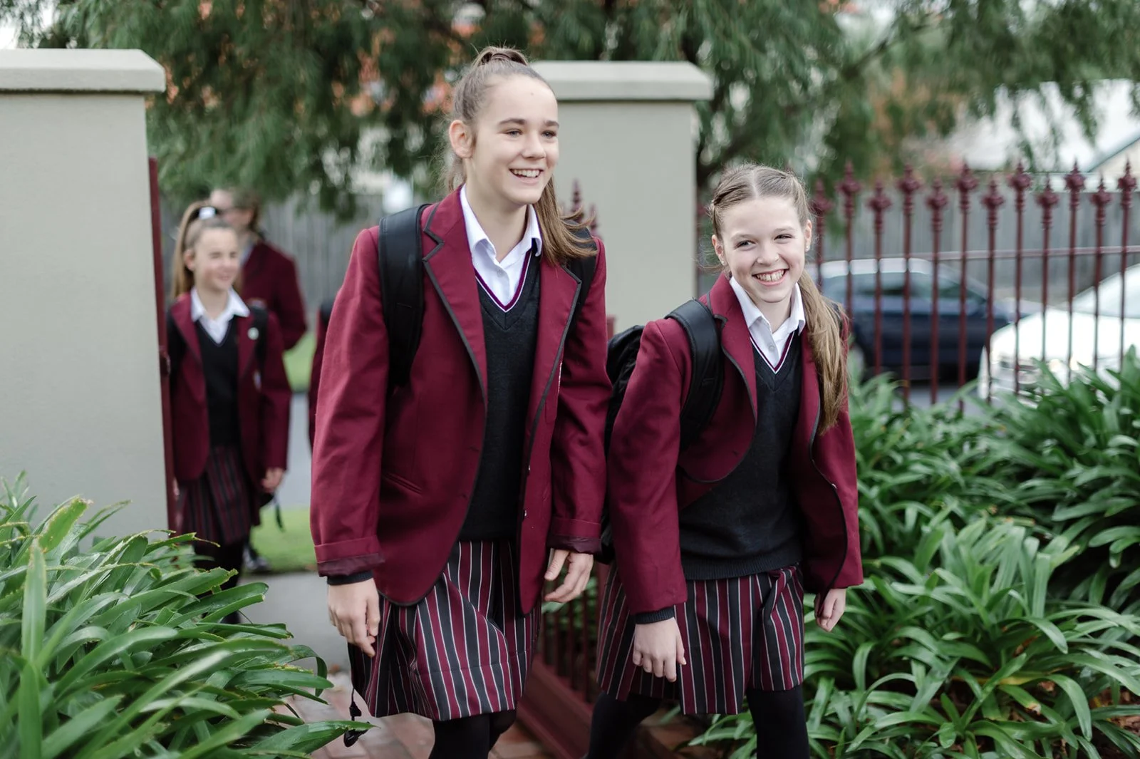 Three schoolgirls in maroon blazers, black sweaters, white shirts, striped skirts, and backpacks walking outdoors among green plants, smiling and talking.