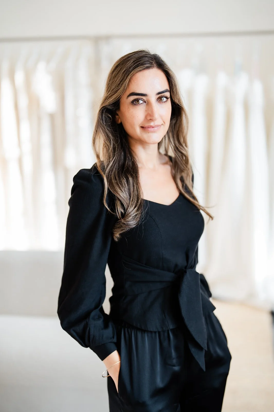 A woman with long wavy hair wearing a black dress with a tied waist, standing indoors with cream-colored curtains in the background.