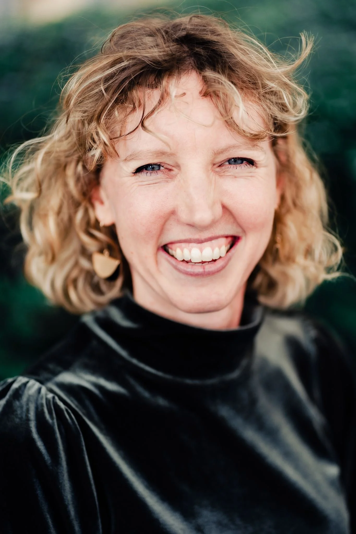 Close-up of a woman with curly blonde hair smiling and squinting outdoors, wearing a black shiny jacket.
