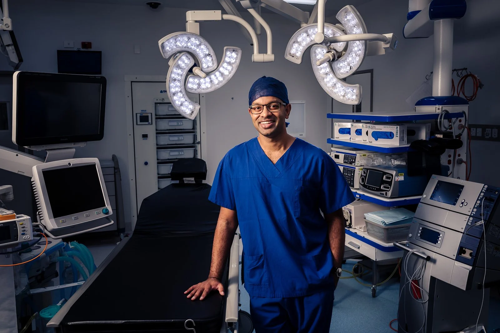A male doctor in scrubs and a surgical cap standing in an operating room, surrounded by medical equipment and surgical lights.
